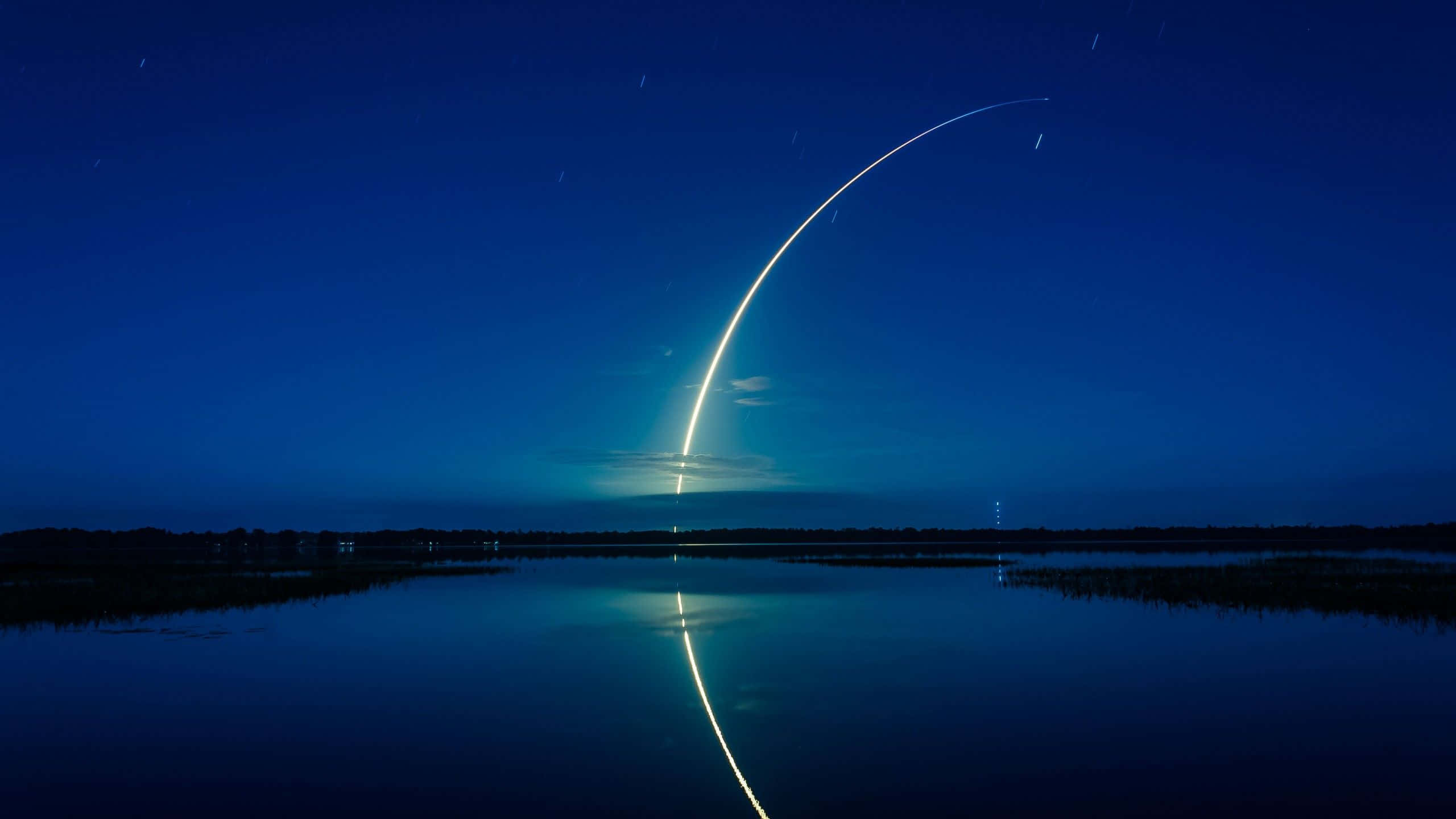 桌布 壁紙 — 在星空下，一枚火箭在夜間升空，留下發光的彎曲軌跡，倒映在平靜的深藍色湖面上。