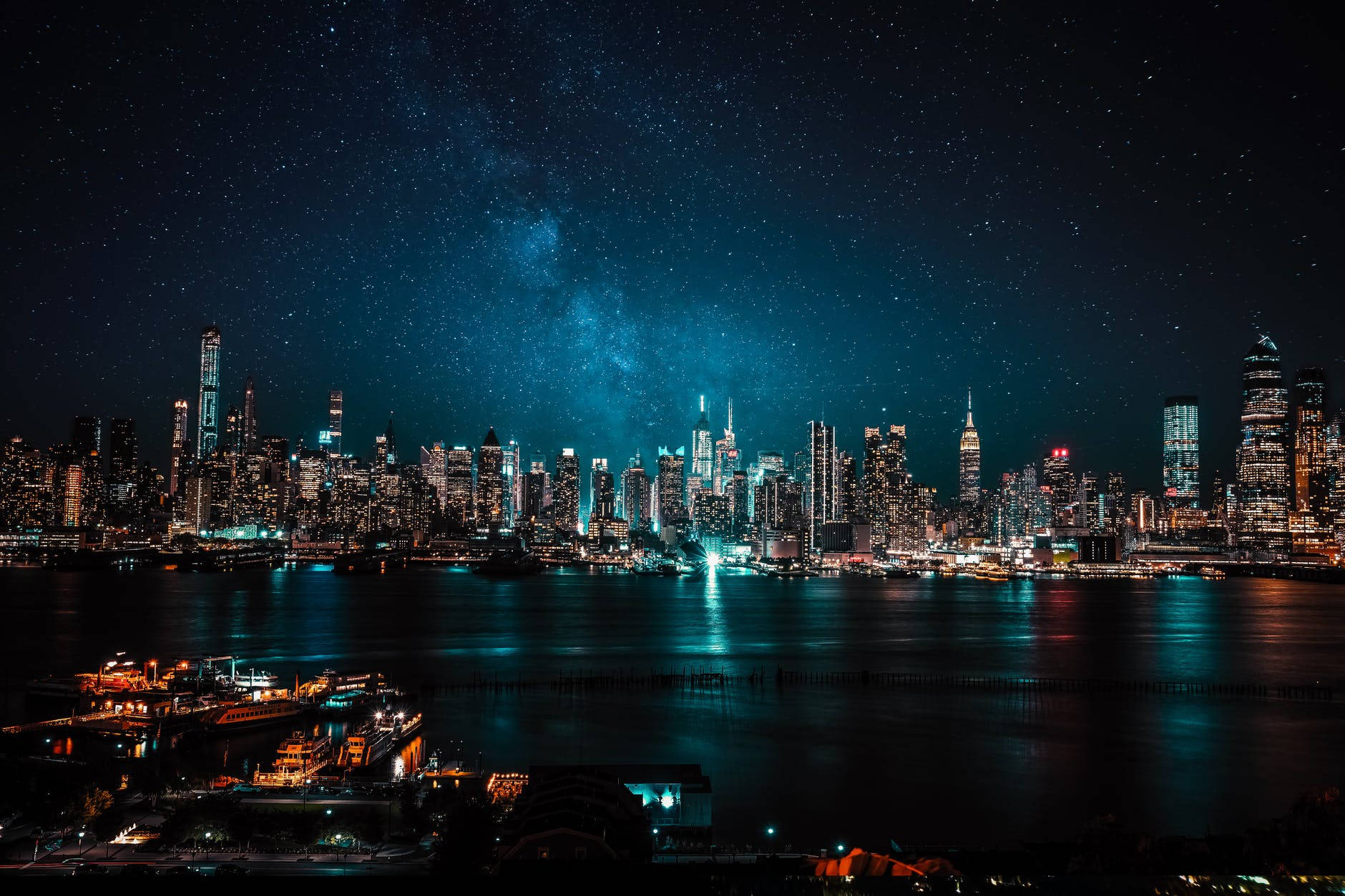 City At Night 桌布 壁紙 — 在星空下，閃爍的摩天大樓和反射的水面上方，銀河拱橋橫跨的紐約天際線夜景，令人目眩神迷。