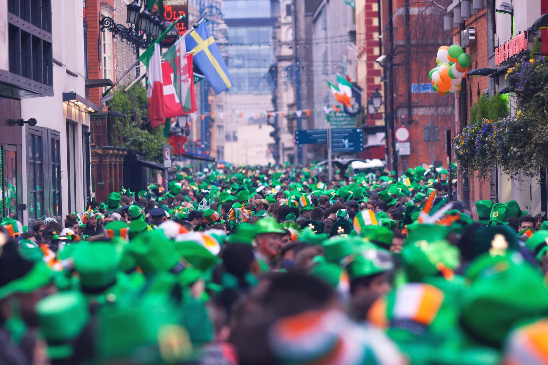 St Patrick's Day 桌布 壁紙 — 在晴朗的天空下，充滿戴著綠色帽子和揮舞愛爾蘭旗幟的人群的熱鬧街頭遊行。