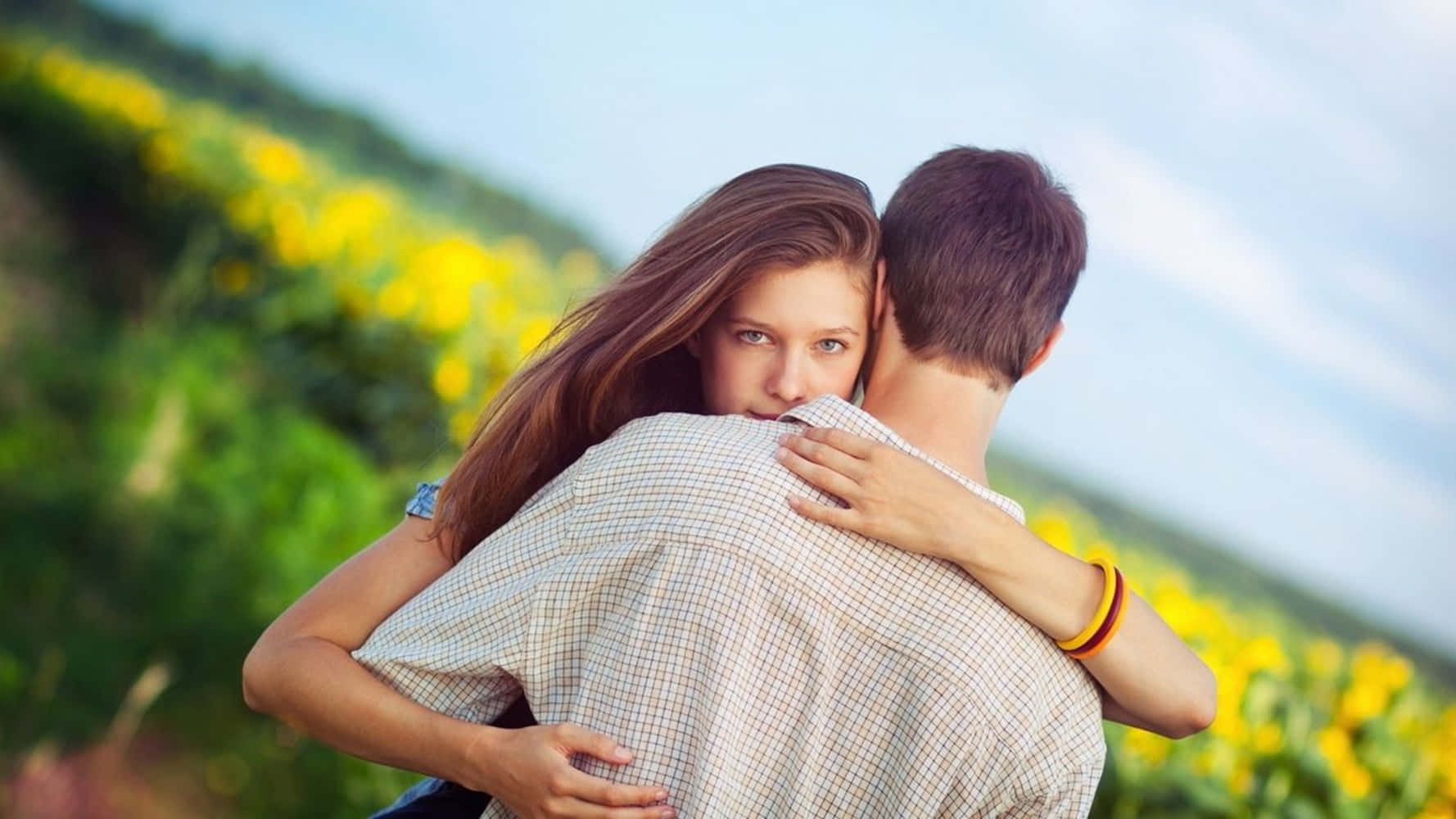 couple love 桌布 壁紙 — 在晴朗的藍天下，一個女人在陽光明媚的黃色花田裡從後面擁抱一個男人。