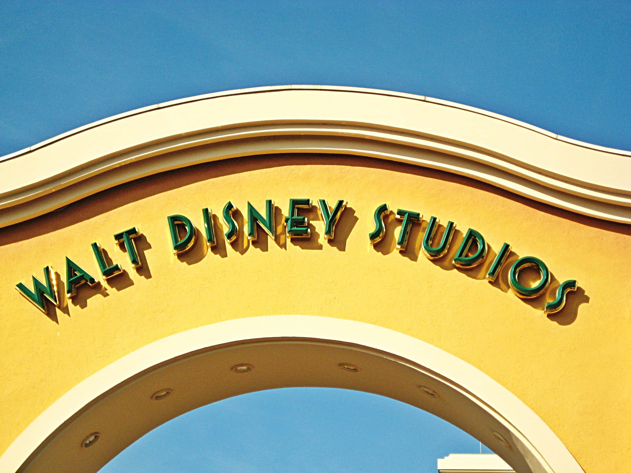 Walt Disney Studios Motion 桌布 壁紙 — 在晴朗的藍天下，一個明亮的黃色拱門上有綠色的「Walt Disney Studios」字母。