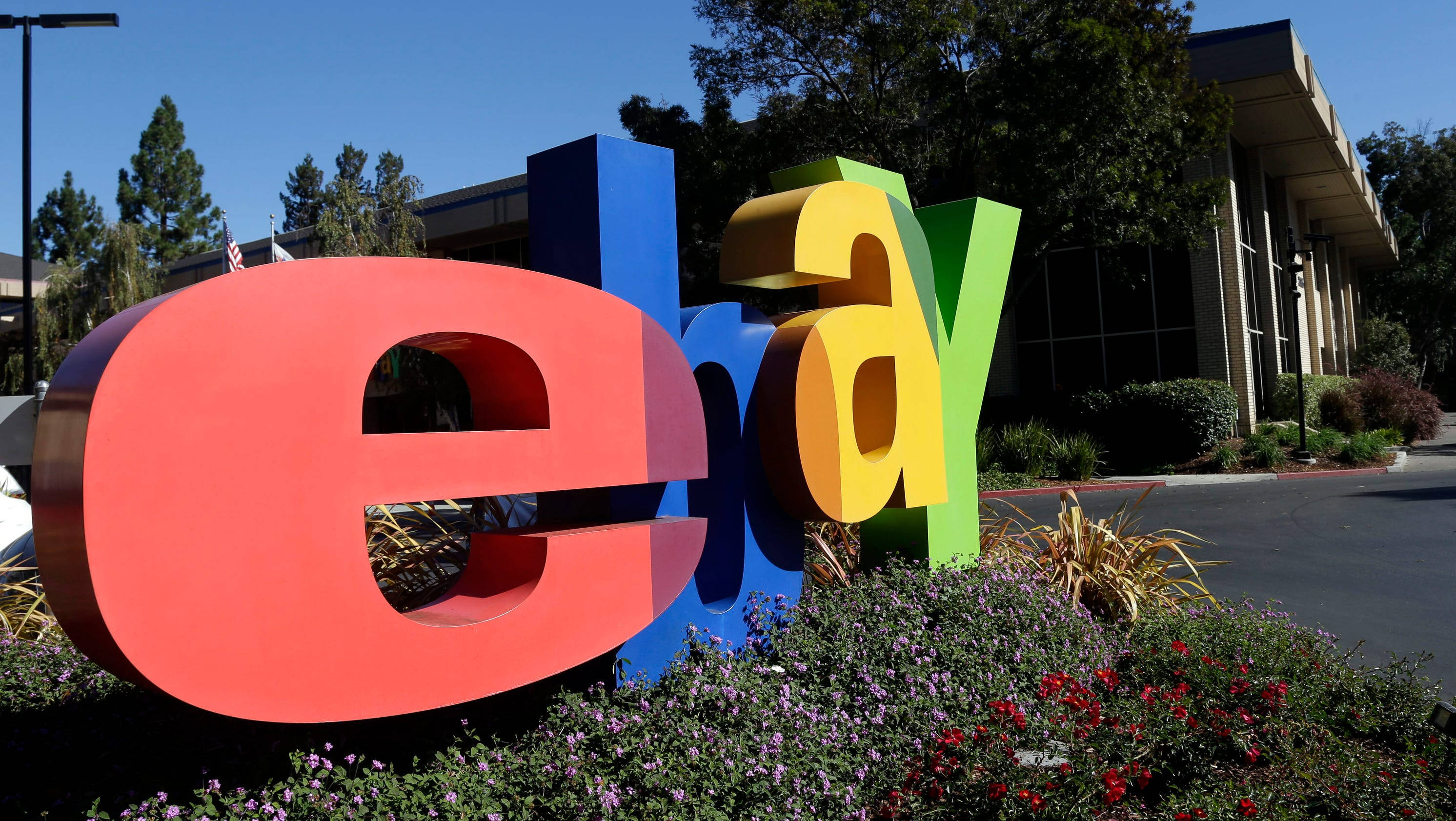 eBay 桌布 壁紙 — 在晴朗的藍天下，一座被開花灌木環繞的建築物前，放置著大型彩色eBay標誌字母。