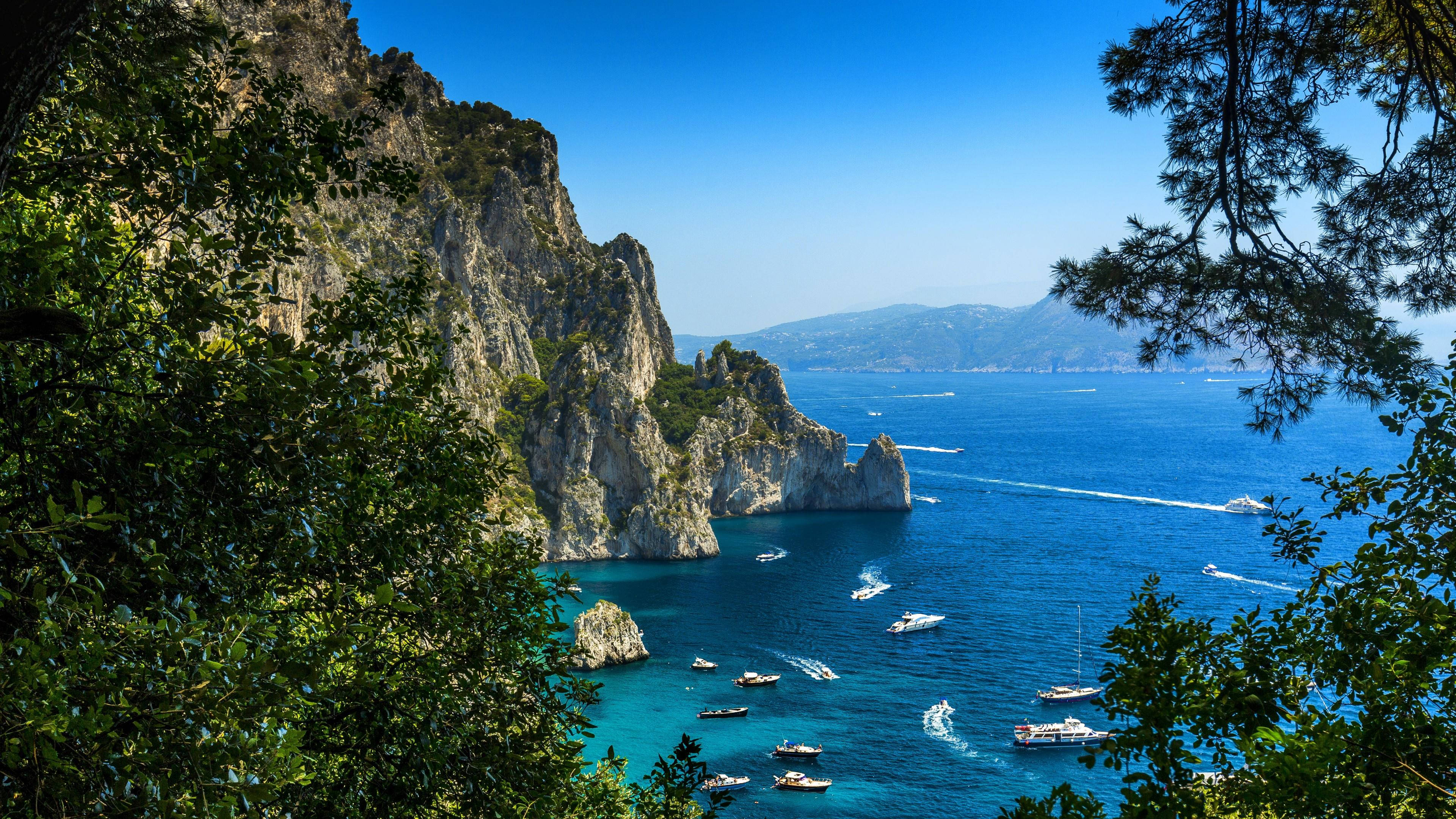 Capri 桌布 壁紙 — 在晴朗的藍天下，卡布里島壯麗的懸崖和碧綠的海水被茂密的綠葉環繞，景色令人嘆為觀止。