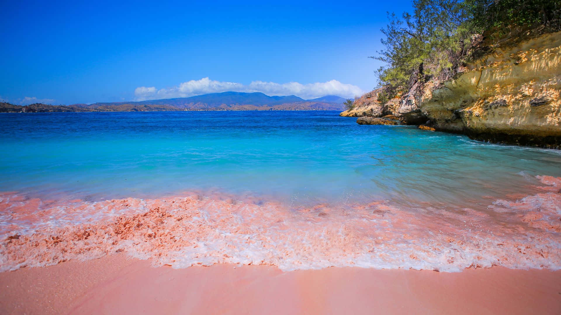 Pink Beach 桌布 壁紙 — 在晴朗的藍天下，碧綠色的海浪拍打著充滿活力的粉紅色沙灘，遠山依稀可見。