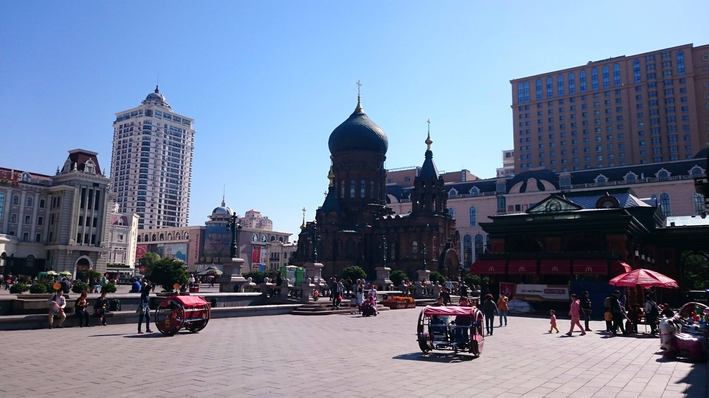 Harbin 桌布 壁紙 — 在晴朗藍天下，一個繁華的城市廣場上有一座引人注目的紅磚大教堂，金色圓頂，周圍環繞著現代摩天大樓和行人。