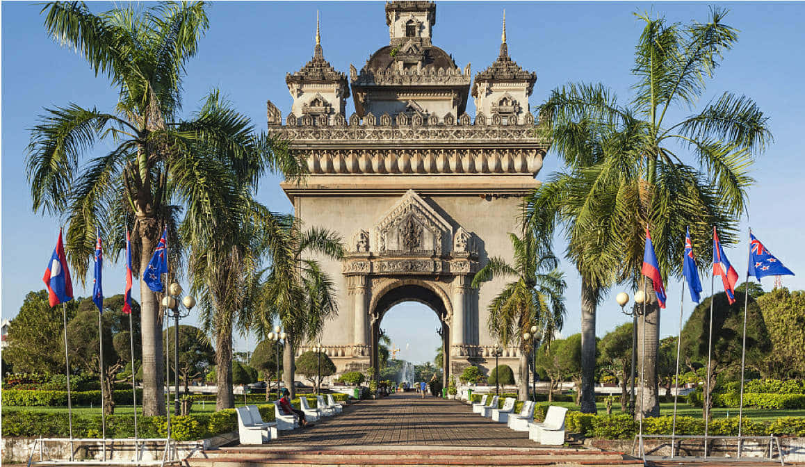 Laos 桌布 壁紙 — 在晴朗藍天下，宏偉華麗的石拱門兩旁種有棕櫚樹並懸掛國旗。