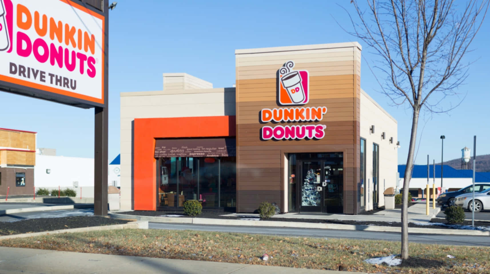 Dunkin Donuts 桌布 壁紙 — 在晴朗藍天下，明亮的橙色和米色Dunkin' Donuts店面，有光禿禿的樹木和停放的汽車。