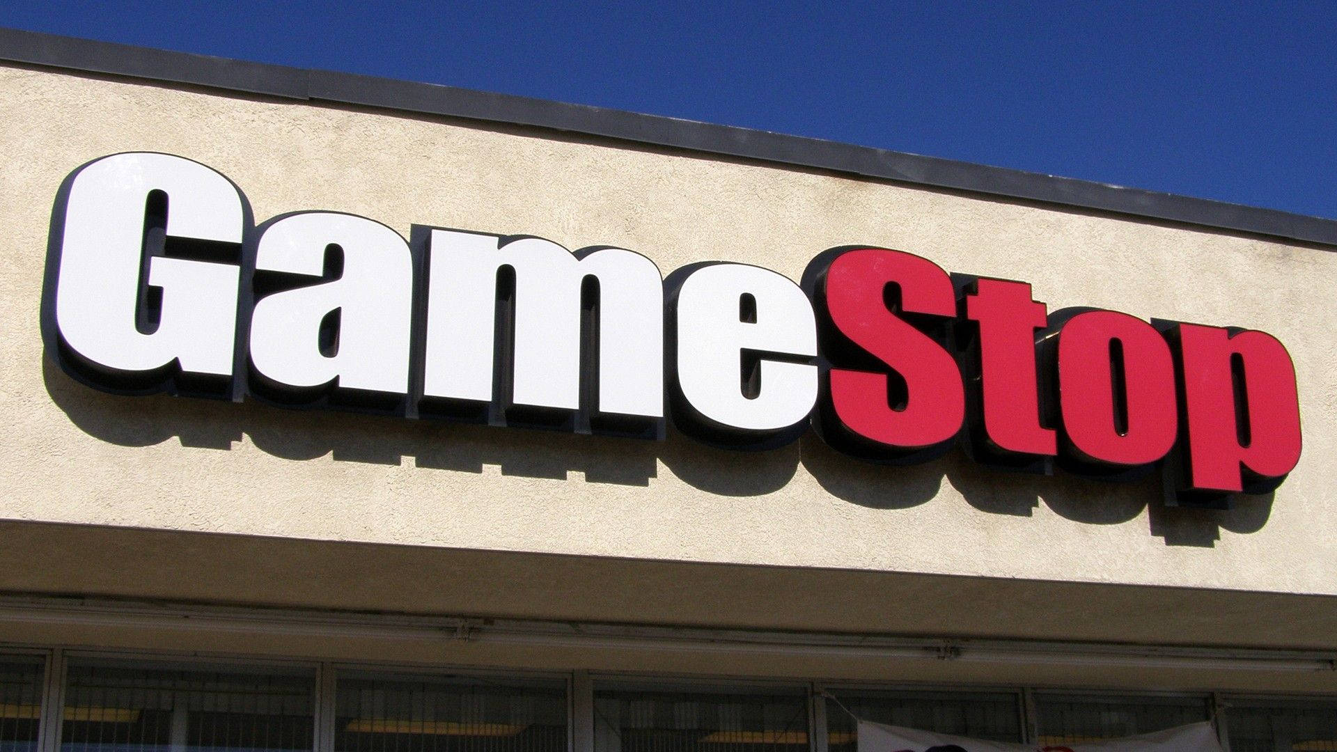 GameStop 桌布 壁紙 — 在晴朗藍天下，米色建築物上安裝了大膽的白色和紅色 GameStop 標誌。