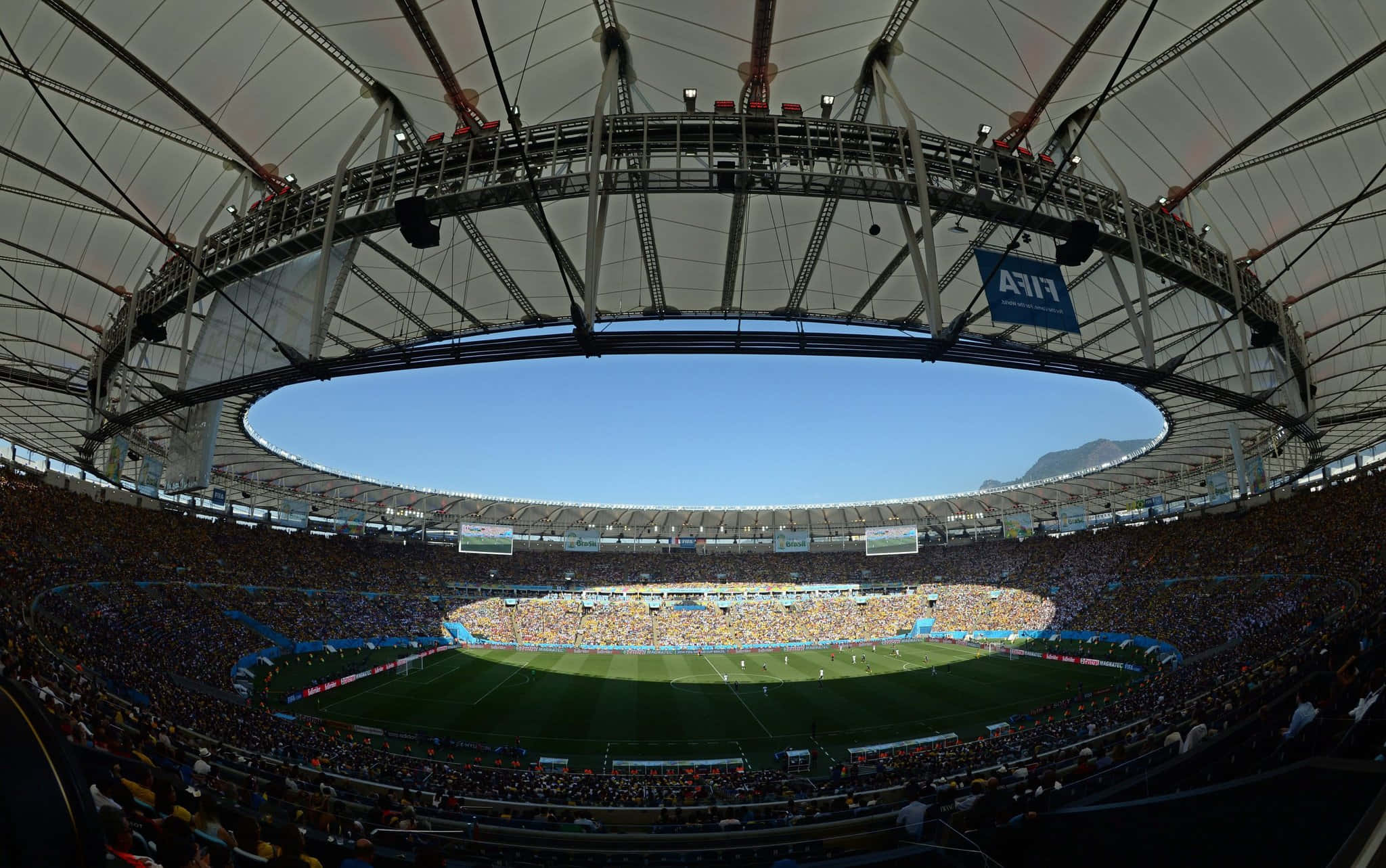 Stadium 桌布 壁紙 — 在晴朗藍天下，遠方有山巒的擠滿觀眾足球場的魚眼視角。