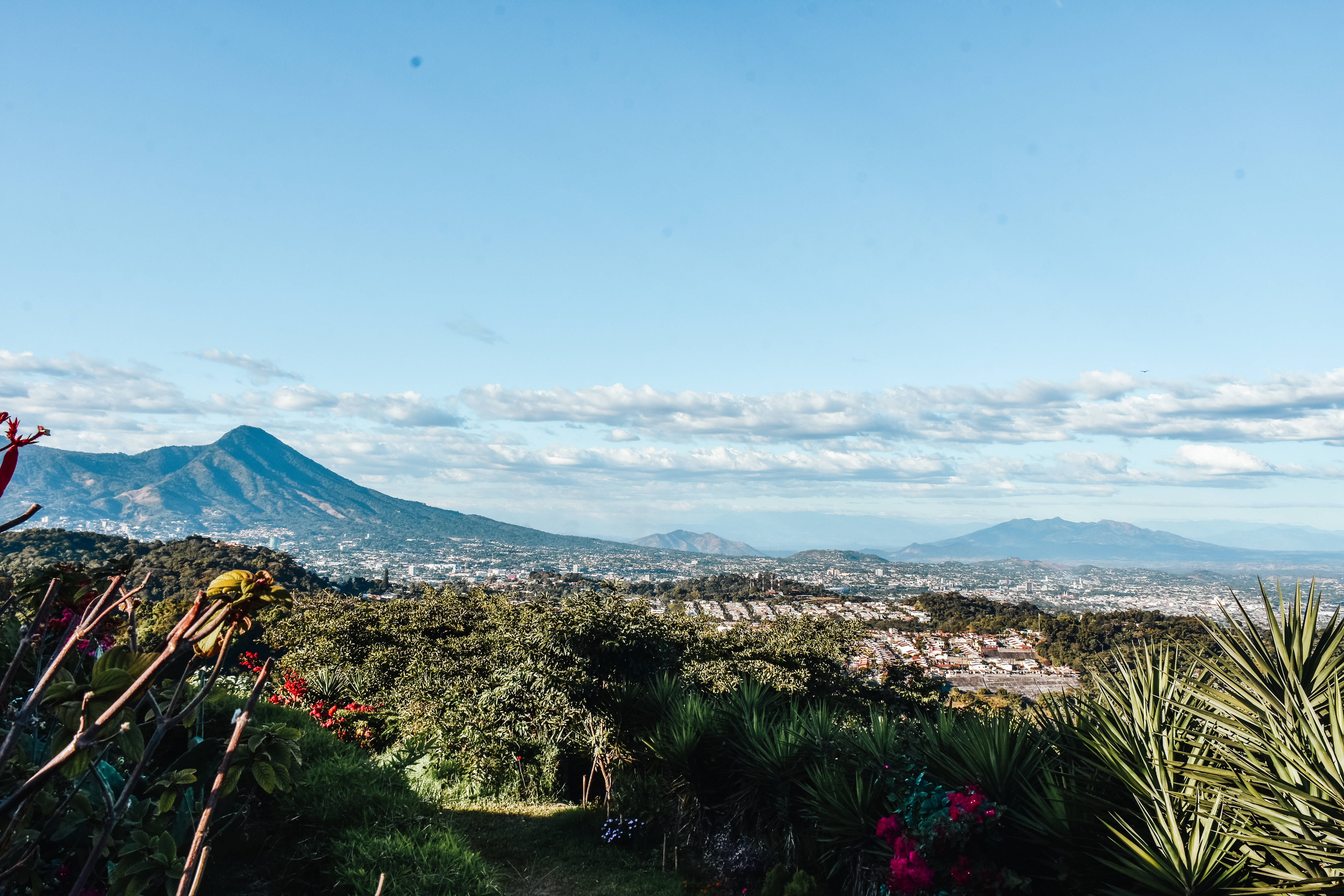 El Salvador 桌布 壁紙 — 在晴朗藍天和零星白雲下，一座宏偉山腳下的廣闊城市全景。