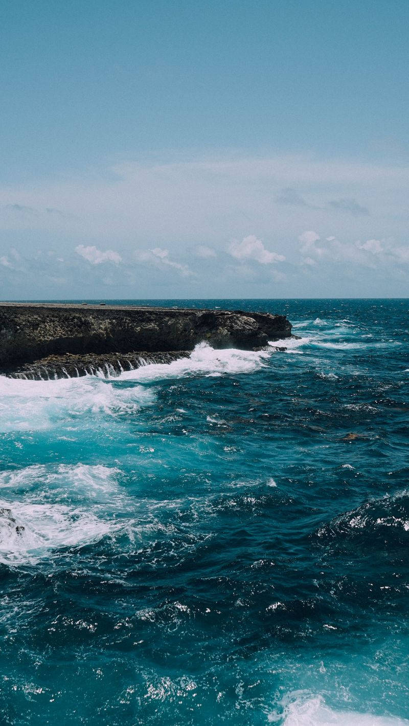 Sea 桌布 壁紙 — 在晴朗藍天和零星白雲下，強勁的藍綠色海浪拍打著崎嶇的黑色懸崖。