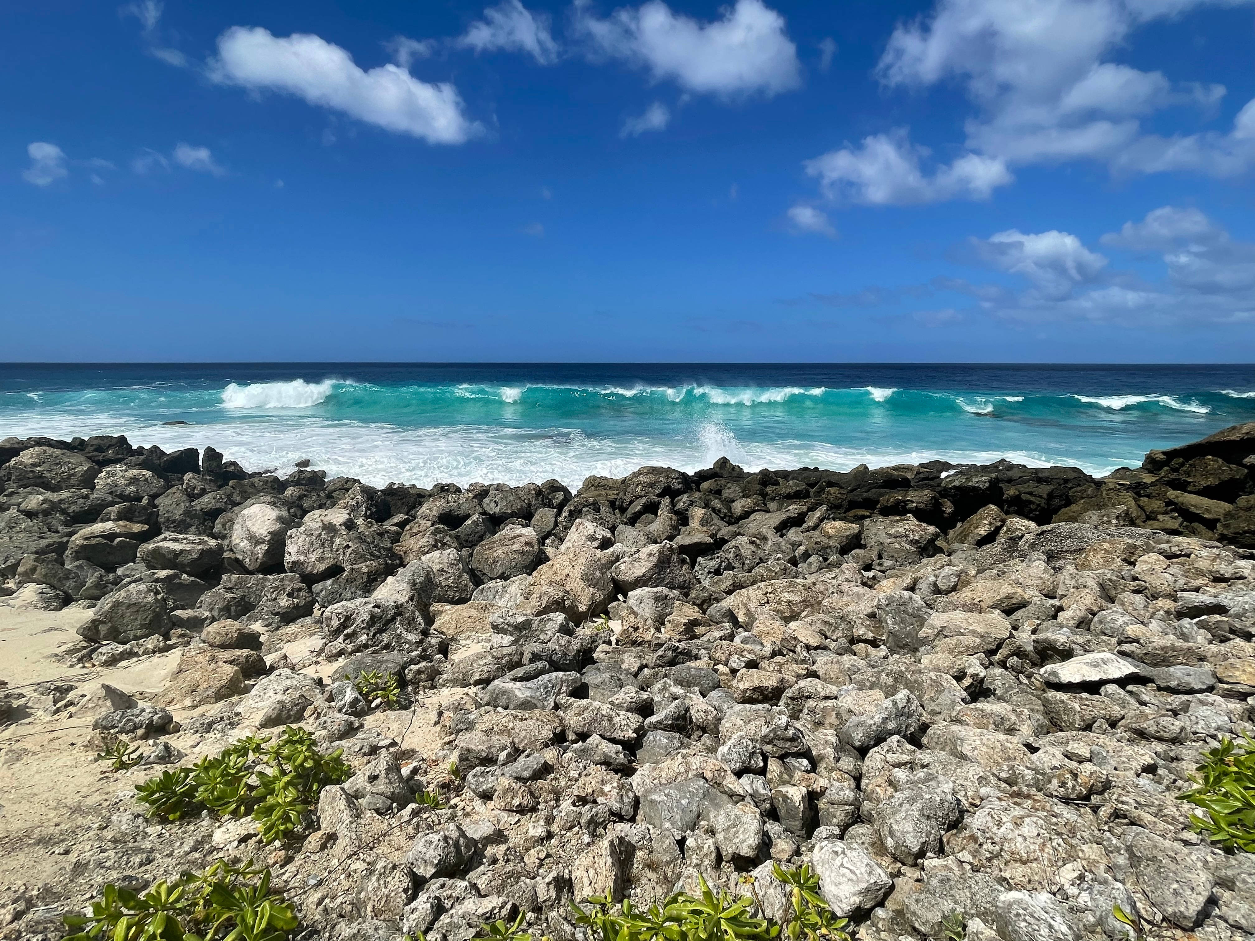 Guam 桌布 壁紙 — 在晴朗藍天和零星白雲下，藍綠色海浪拍打著粗獷的火山岩前景。