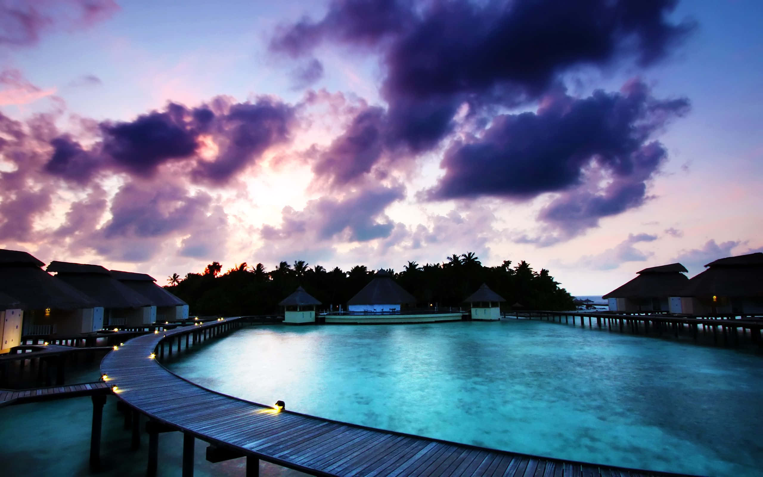 Bora Bora 桌布 壁紙 — 在暮色下，一條木製步道蜿蜒穿過碧綠色的潟湖，上方是戲劇性的紫色和粉紅色天空。