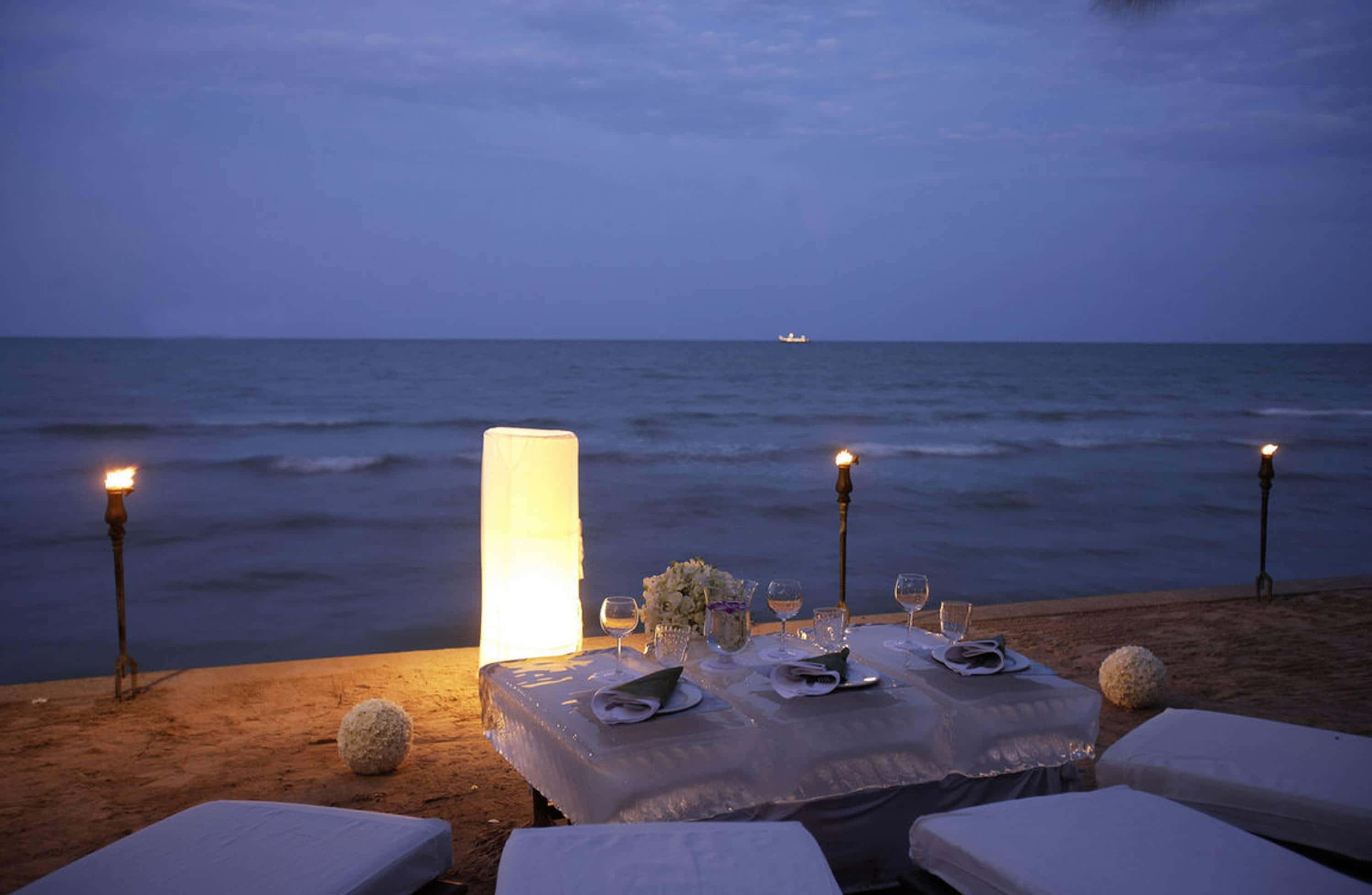 Beach Picnic 桌布 壁紙 — 在暮色中，一張浪漫的海邊餐桌，點綴著發光的燈籠和蠟燭，背景是平靜的海平線。
