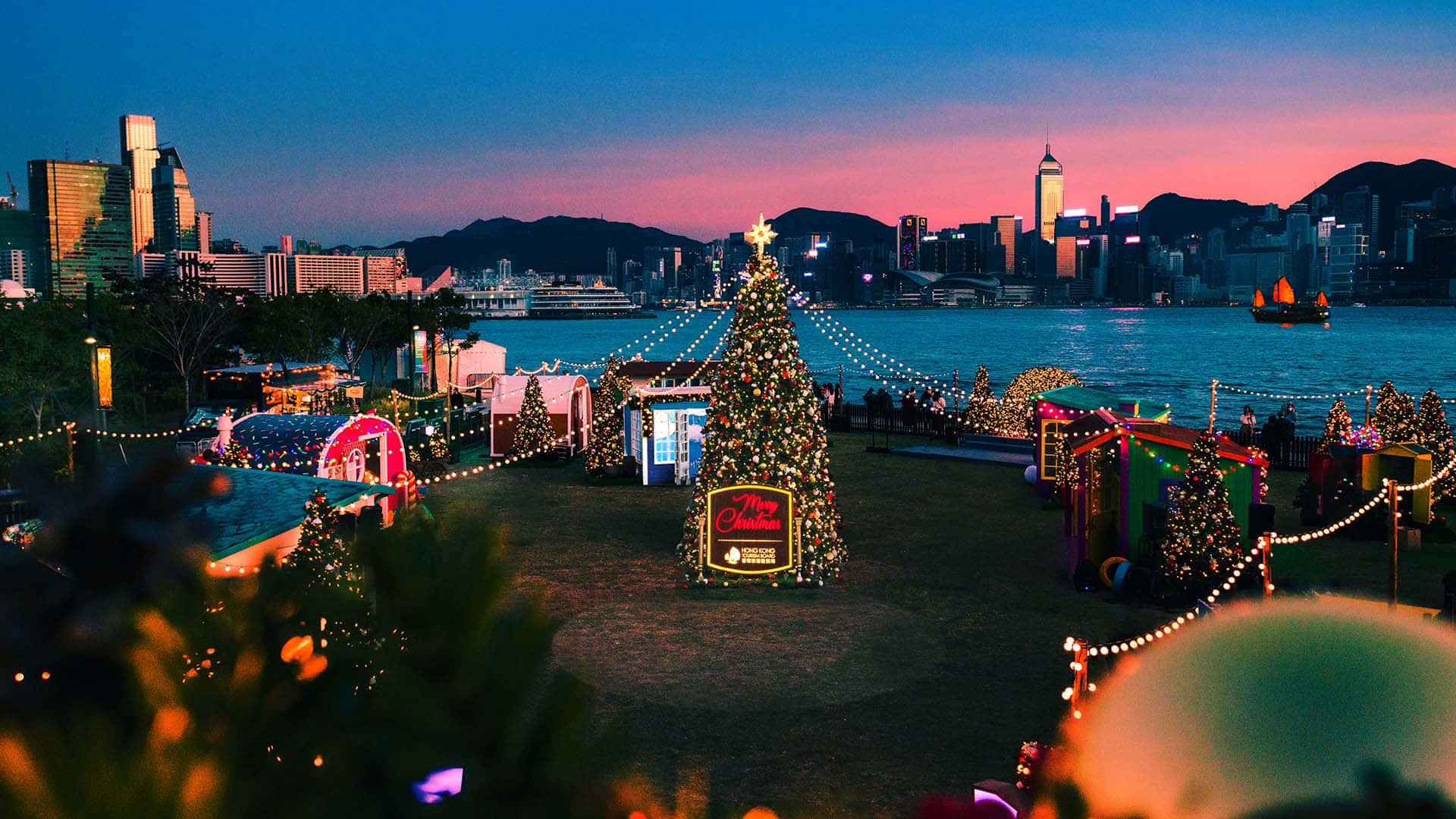 Christmas Village 桌布 壁紙 — 在暮色中，一棵節日聖誕樹在水邊市場旁閃耀，市場有色彩繽紛的攤位，背景是城市天際線。
