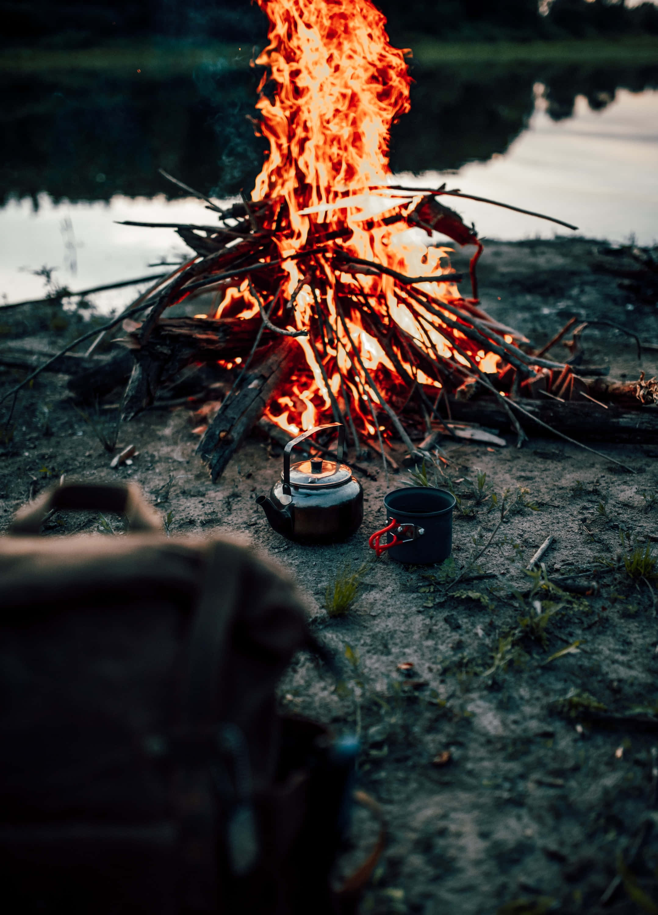 Fall Bonfire 桌布 壁紙 — 在暮色天空下，一個咆哮的營火在平靜的湖邊明亮地燃燒，附近放著水壺和馬克杯。