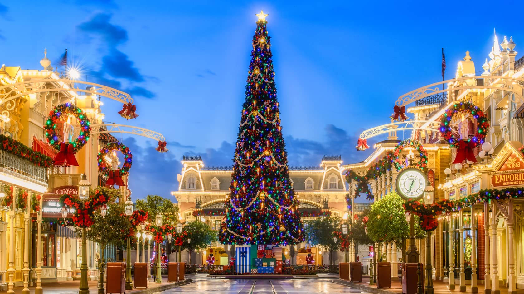 Christmas Tree 桌布 壁紙 — 在暮色天空下，一棵大型亮燈聖誕樹矗立在充滿節日氣氛的迪士尼風格城鎮廣場中央，廣場裝飾著花環和閃爍的燈光。