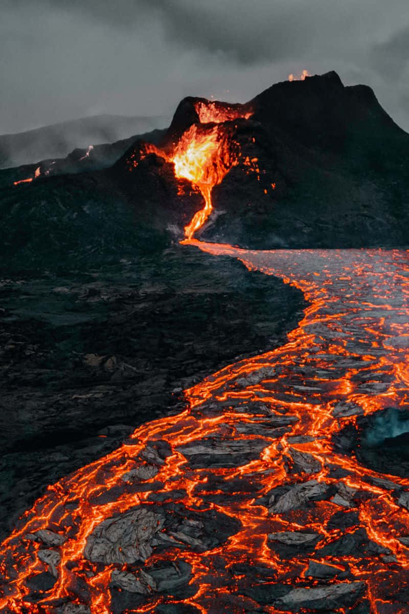 lava 桌布 壁紙 — 在暴風雨灰濛濛的天空下，一條熾熱的橙色熔岩河流從黑暗的火山山上傾瀉而下。