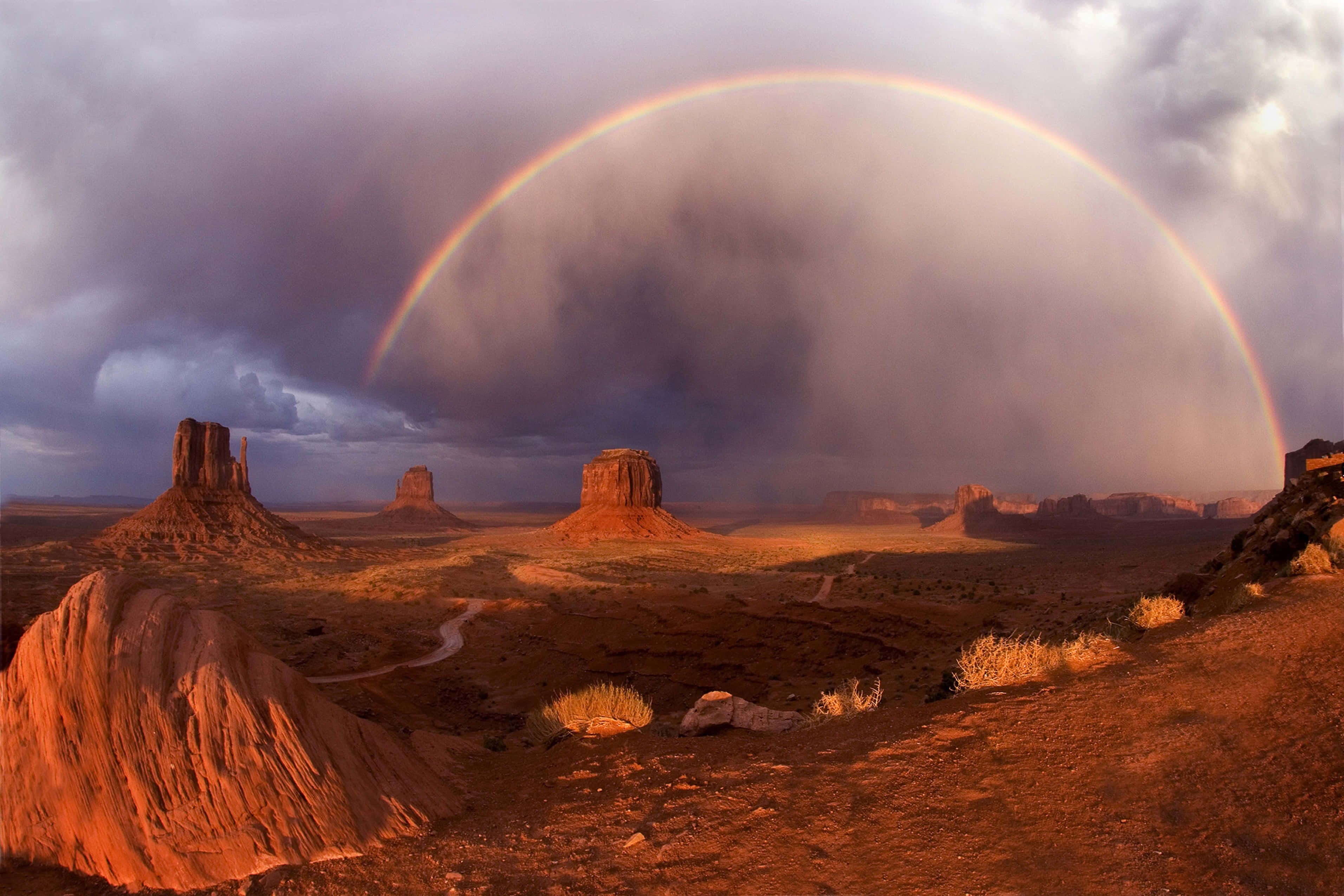 Arizona 桌布 壁紙 — 在暴風雨的天空下，一道鮮豔的彩虹在沙漠景觀中戲劇性的紅色岩層上拱起。