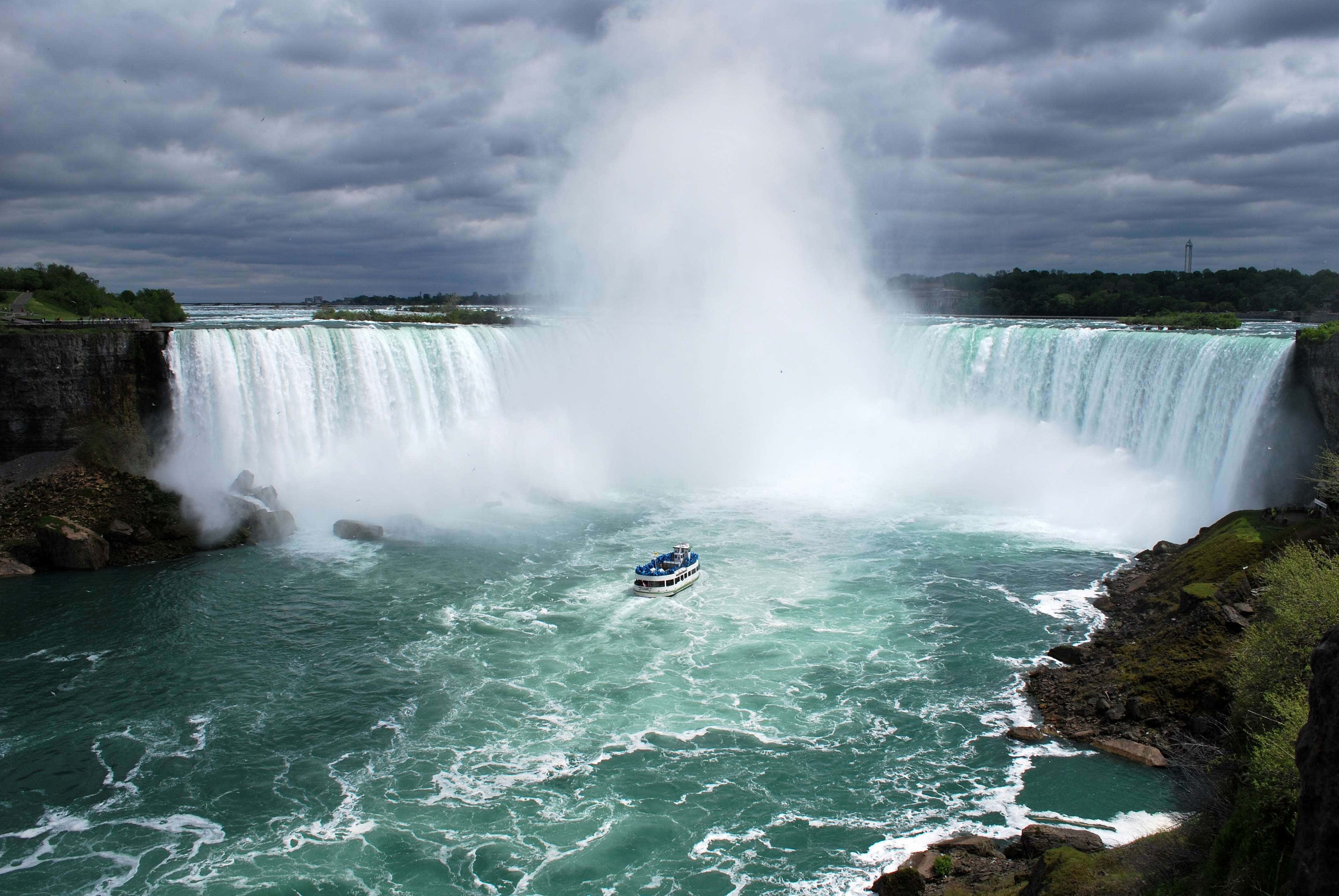 720p Niagara Falls 桌布 壁紙 — 在暴風雨的天空下，尼亞加拉大瀑布的雄偉廣角景觀，一艘觀光船在下方洶湧的碧綠海水中航行。