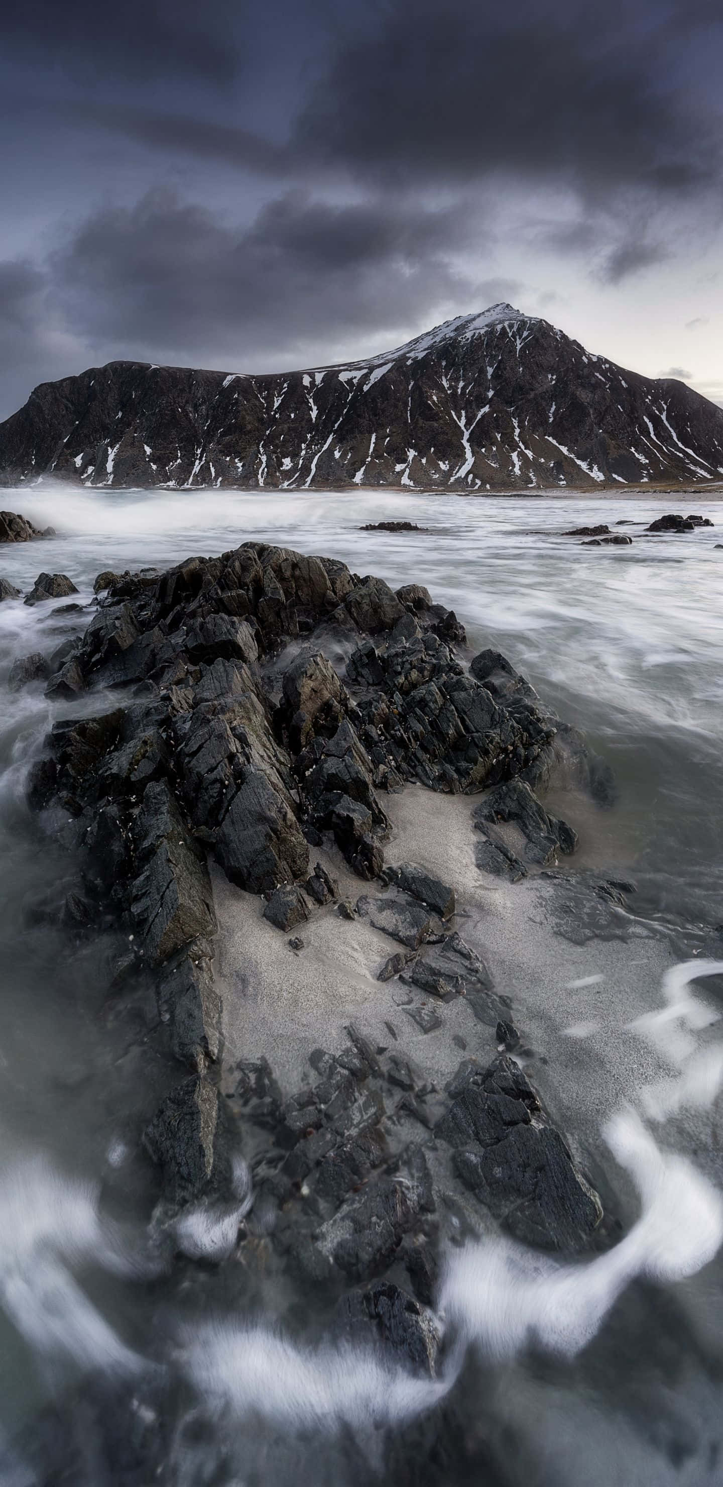 1440x2960 桌布 壁紙 — 在暴風雨的天空下，泡沫般的海浪拍打著黑暗的岩石，遠處有積雪的山脈，這是一幅戲劇性的長時間曝光海景。