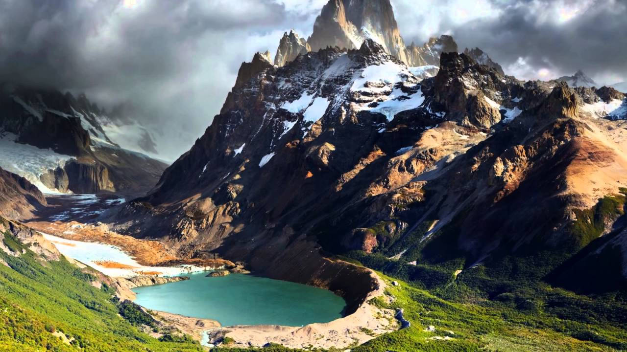 山景 桌布 壁紙 — 在暴風雨的天空下，翠綠山谷中坐落著一座戲劇性的山脈，山頂積雪，湖泊呈藍綠色。