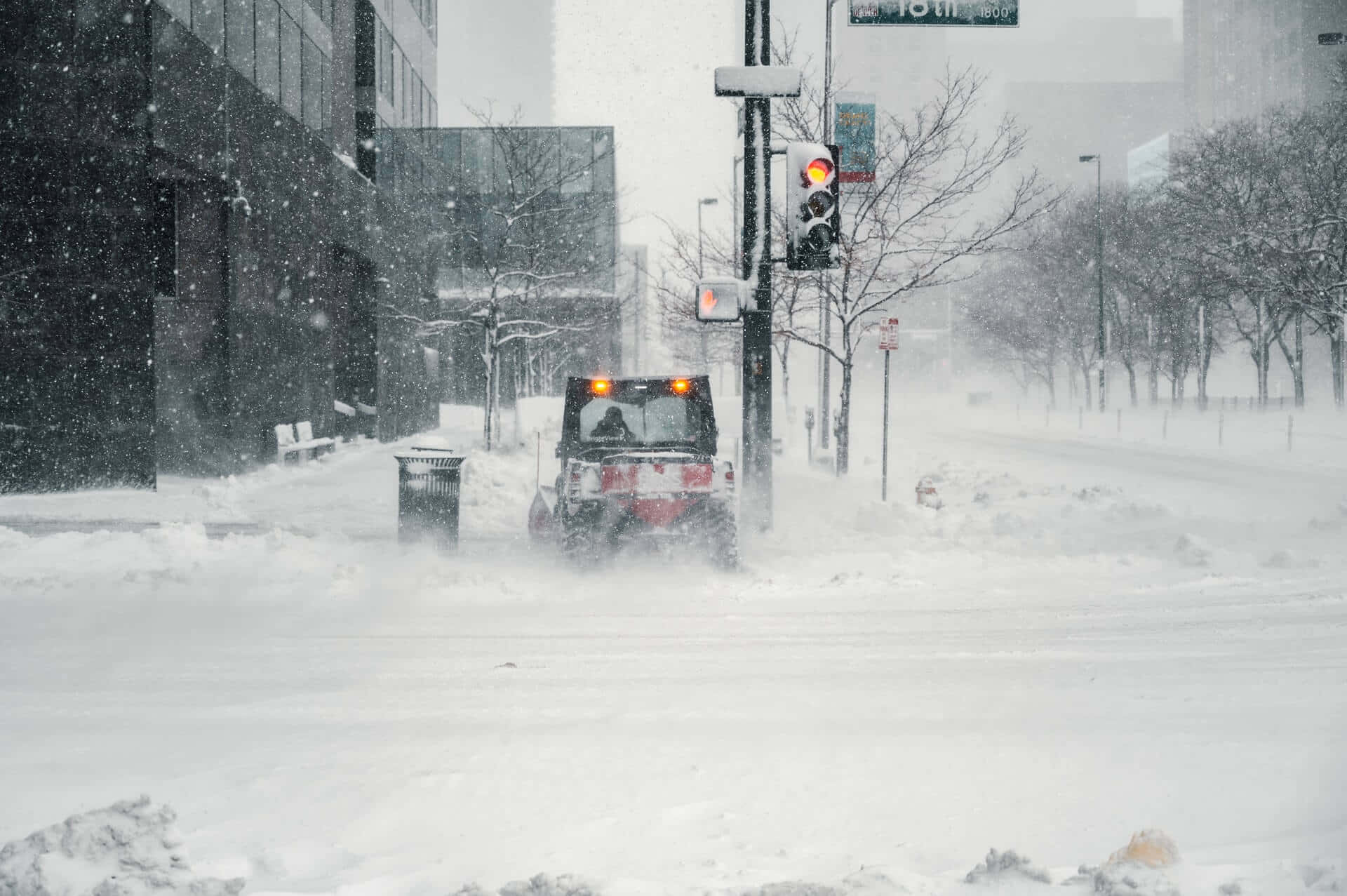 blizzard 桌布 壁紙 — 在暴風雪中，一輛掃雪車正在清理積雪覆蓋的城市街道，雪花飄落，樹木光禿。