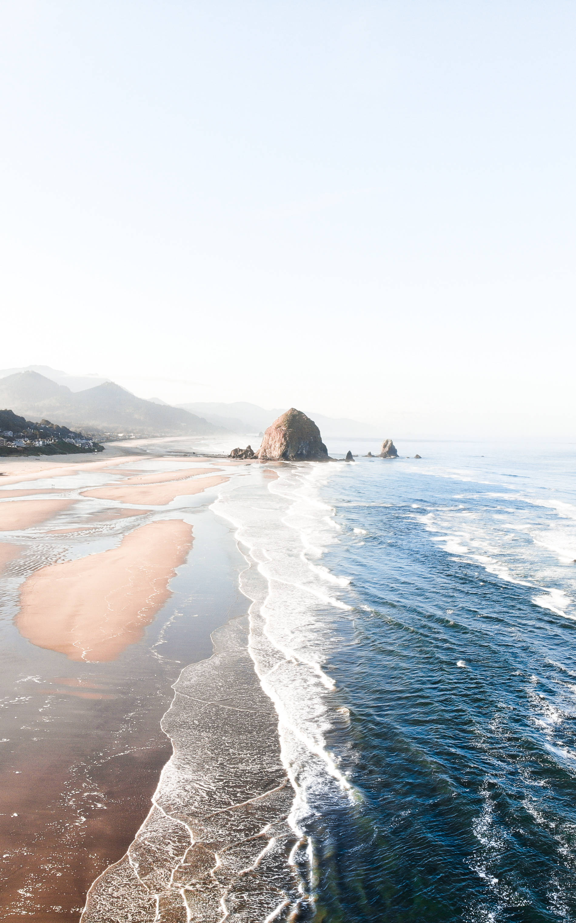 海灘 iPhone 桌布 壁紙 — 在朦朧的天空下，一個寧靜的海岸場景，溫柔的海浪拍打著粉紅色的沙灘，旁邊有一個巨大的海蝕柱。