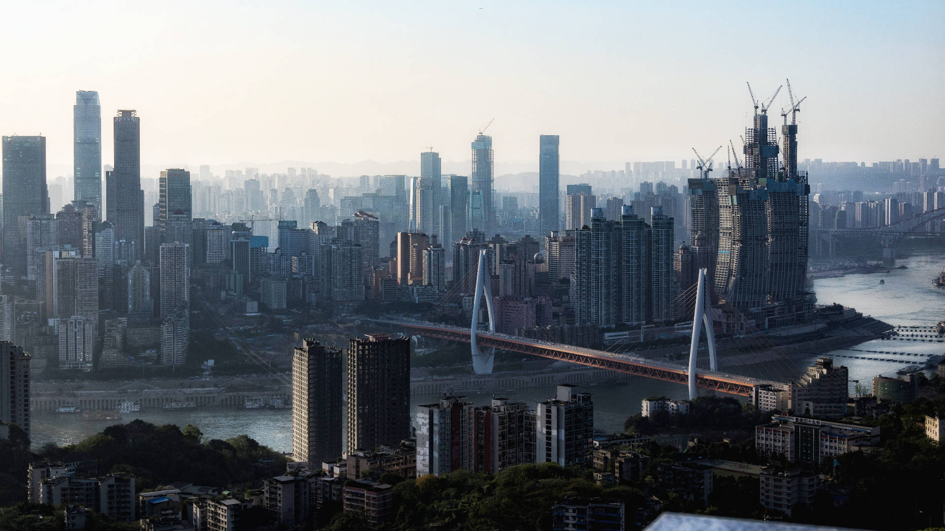 Chongqing 桌布 壁紙 — 在朦朧的日光下，廣闊的城市天際線，高聳的摩天大樓和一條獨特的曲線橋橫跨寬闊的河流。