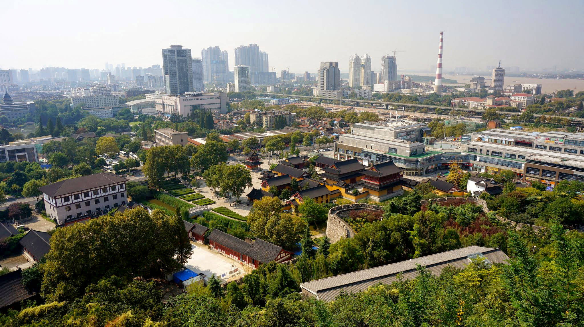 Nanjing 桌布 壁紙 — 在朦朧的日光下，現代城市天際線中鬱鬱蔥蔥的綠樹環繞著的傳統中式寺廟的鳥瞰圖。