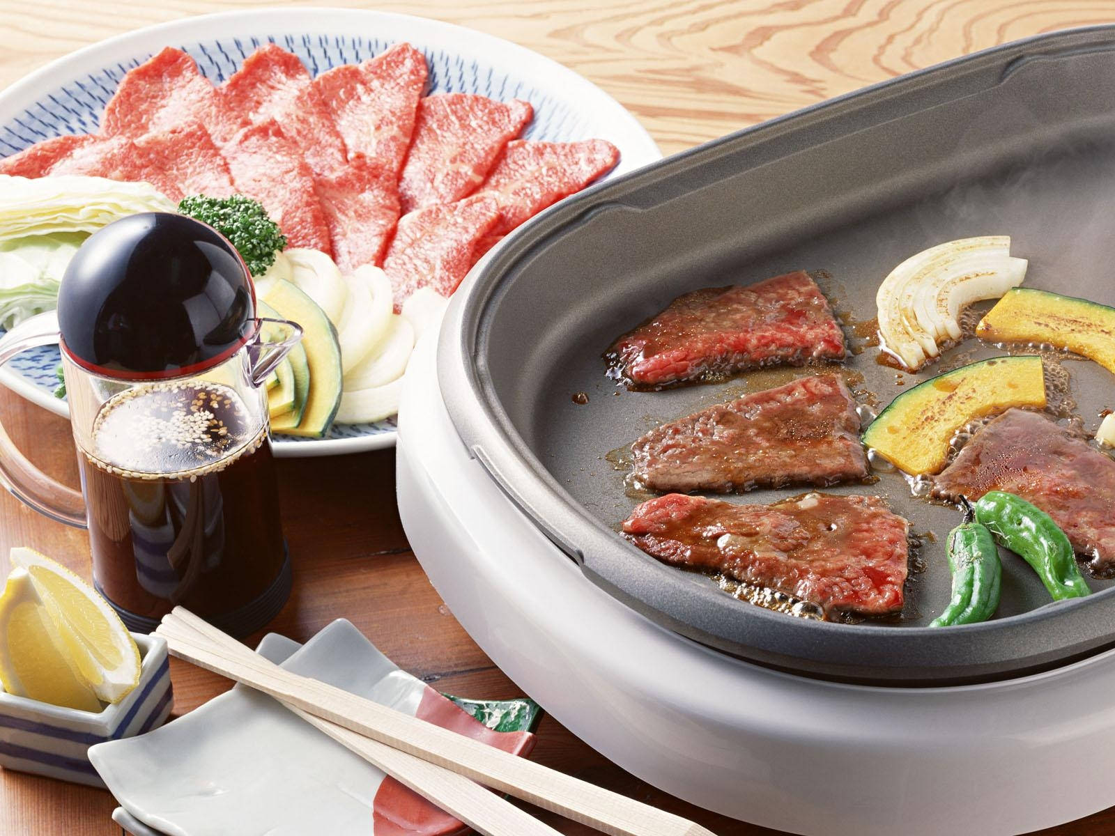 meat 桌布 壁紙 — 在木桌上，熱電烤盤上滋滋作響的牛肉片和蔬菜正在烹調，旁邊放著一盤生牛肉和沾醬。