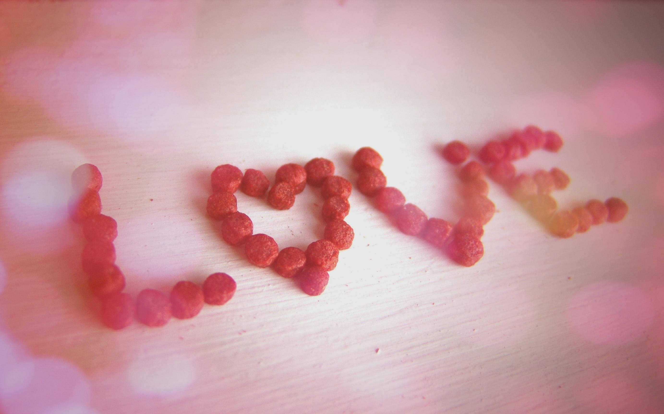 Sweet Love 桌布 壁紙 — 在木質表面上用紅色寵物食品拼寫出「love」一詞，背景為柔和粉紅色散景。