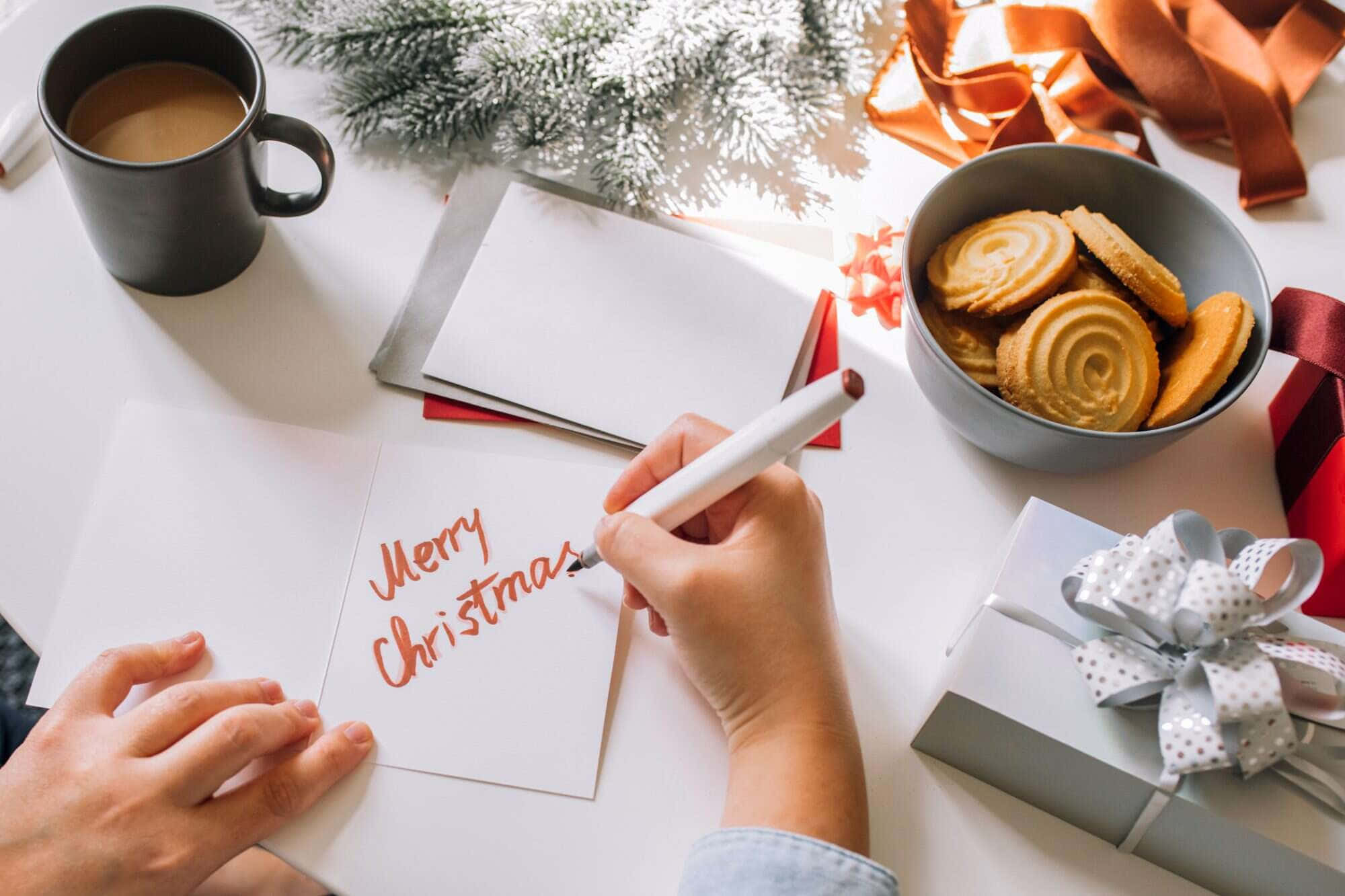 Christmas Card 桌布 壁紙 — 在柔和的冬日燈光下，一隻手在卡片上寫上「聖誕快樂」，旁邊有餅乾、杯子和包裝好的禮物。