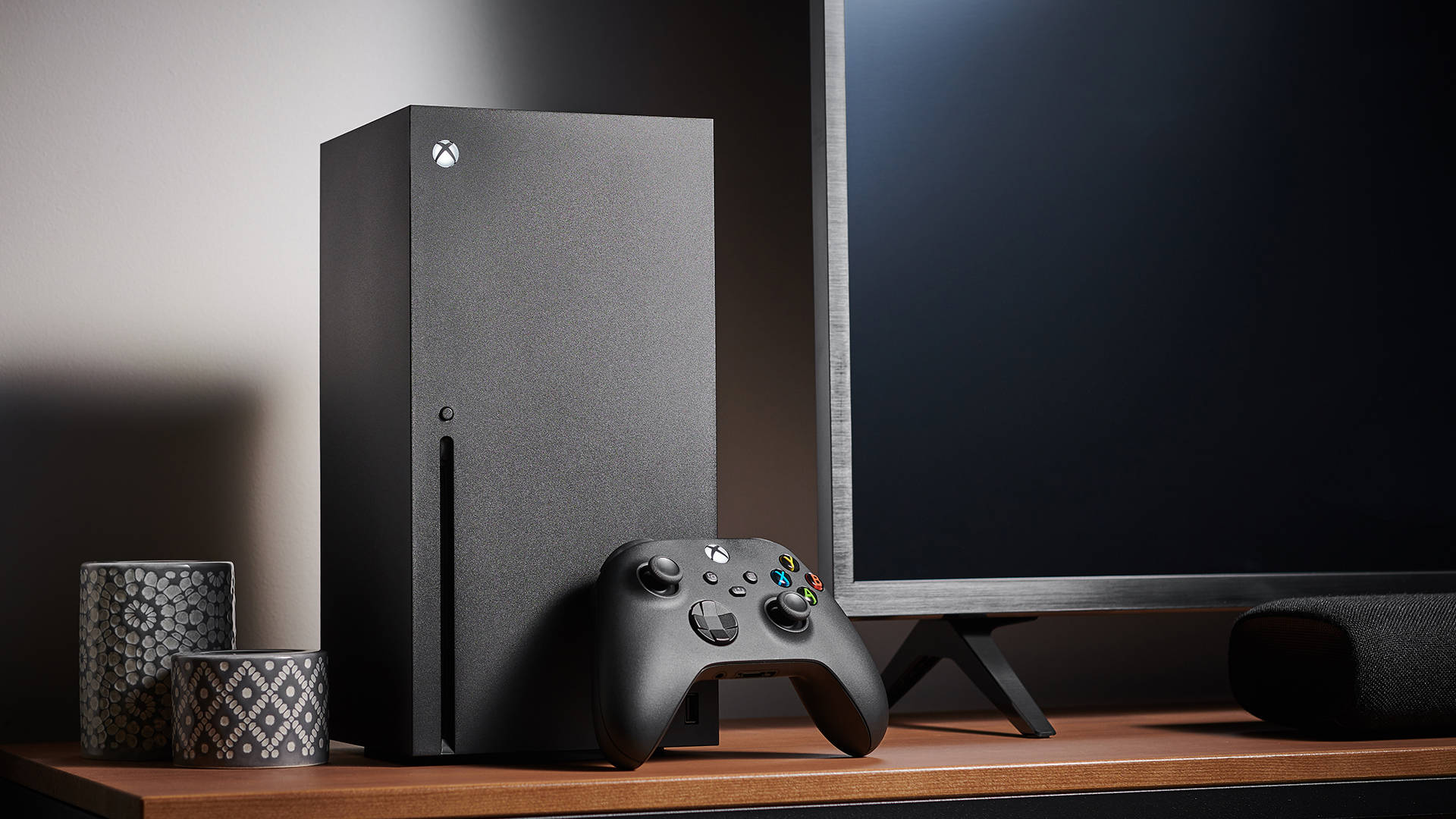 X 桌布 壁紙 — 在柔和的工作室燈光下，一個時尚的黑色 Xbox Series X 主機和控制器放在木架上，旁邊有電視和圖案燭台。