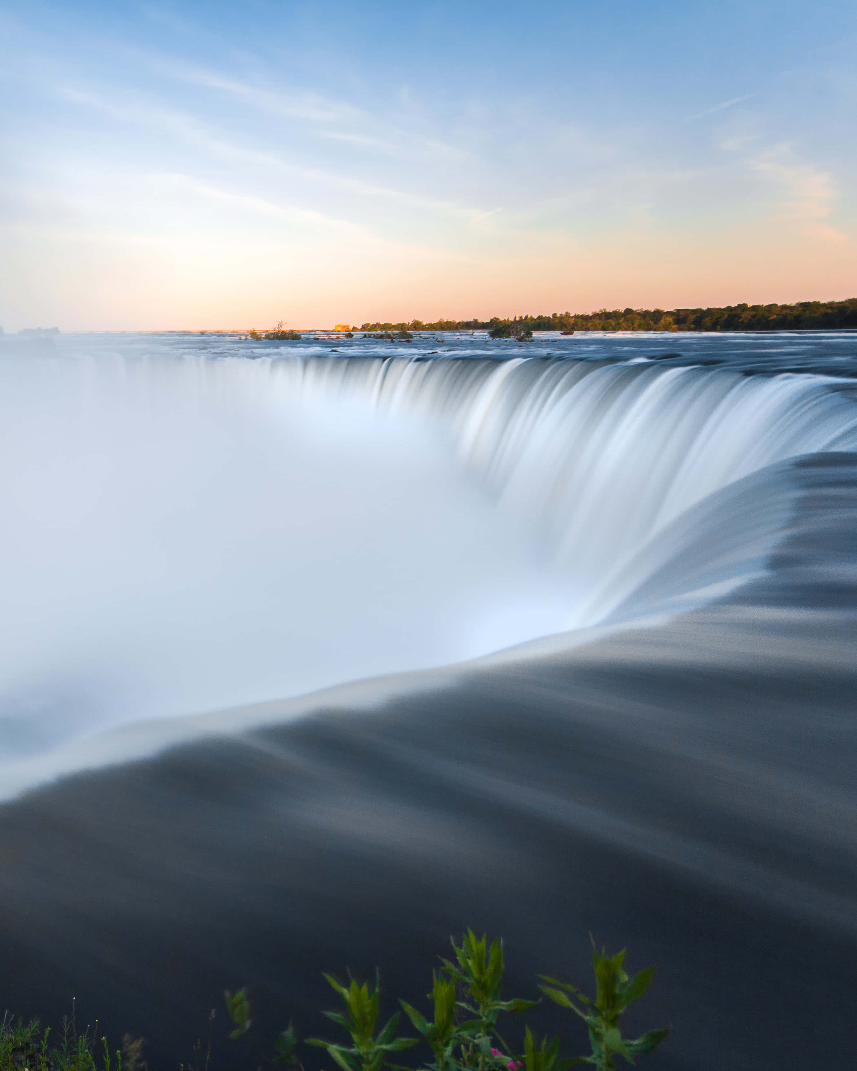 Niagara Falls Canada 桌布 壁紙 — 在柔和的日出天空下，薄雲和綠色樹葉在前景，一個壯麗的瀑布以慢動作傾瀉而下。
