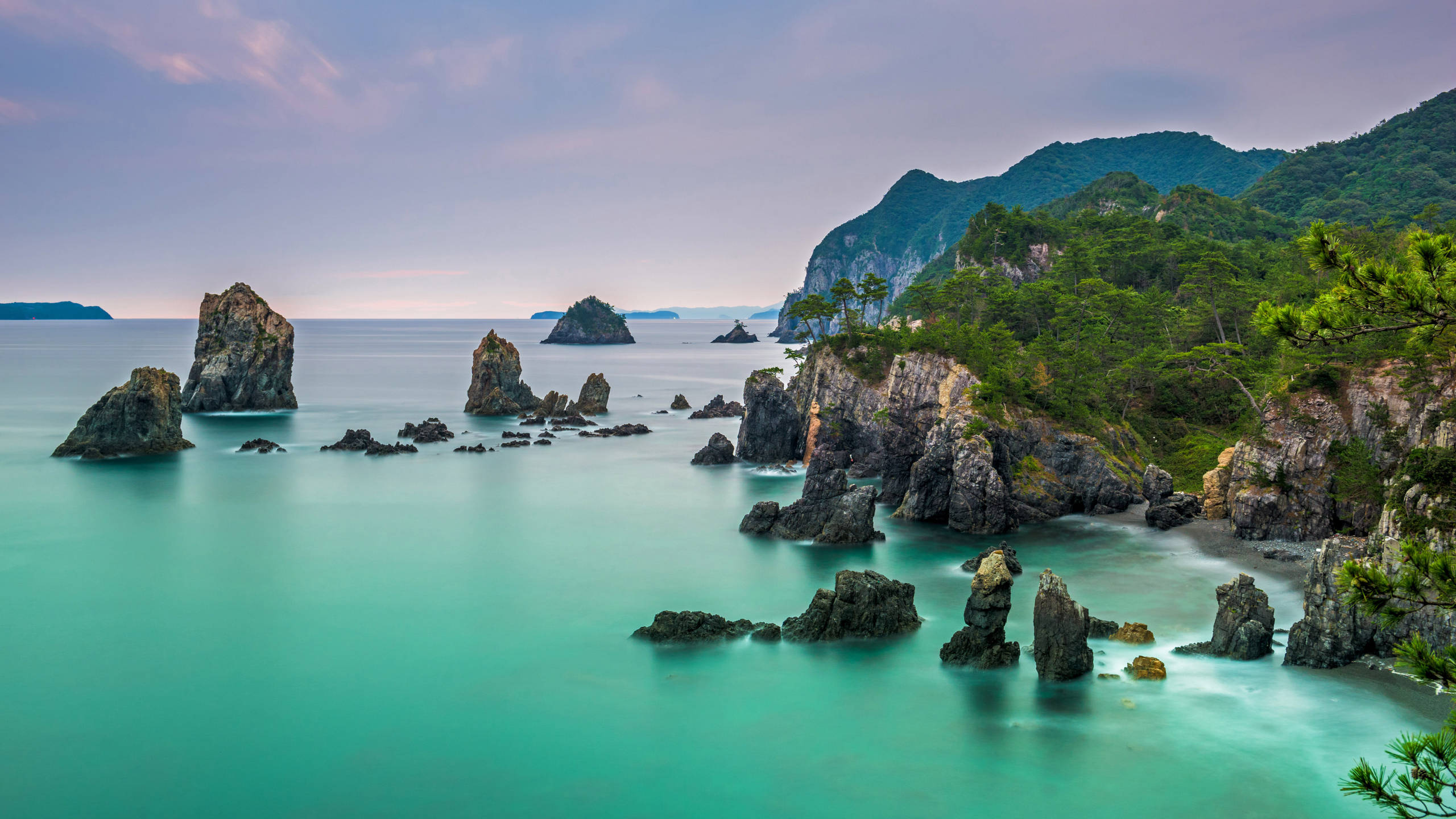 2560x1440 nature 桌布 壁紙 — 在柔和的暮色天空下，碧綠色的海水輕輕拍打著參差不齊的黑色岩石。