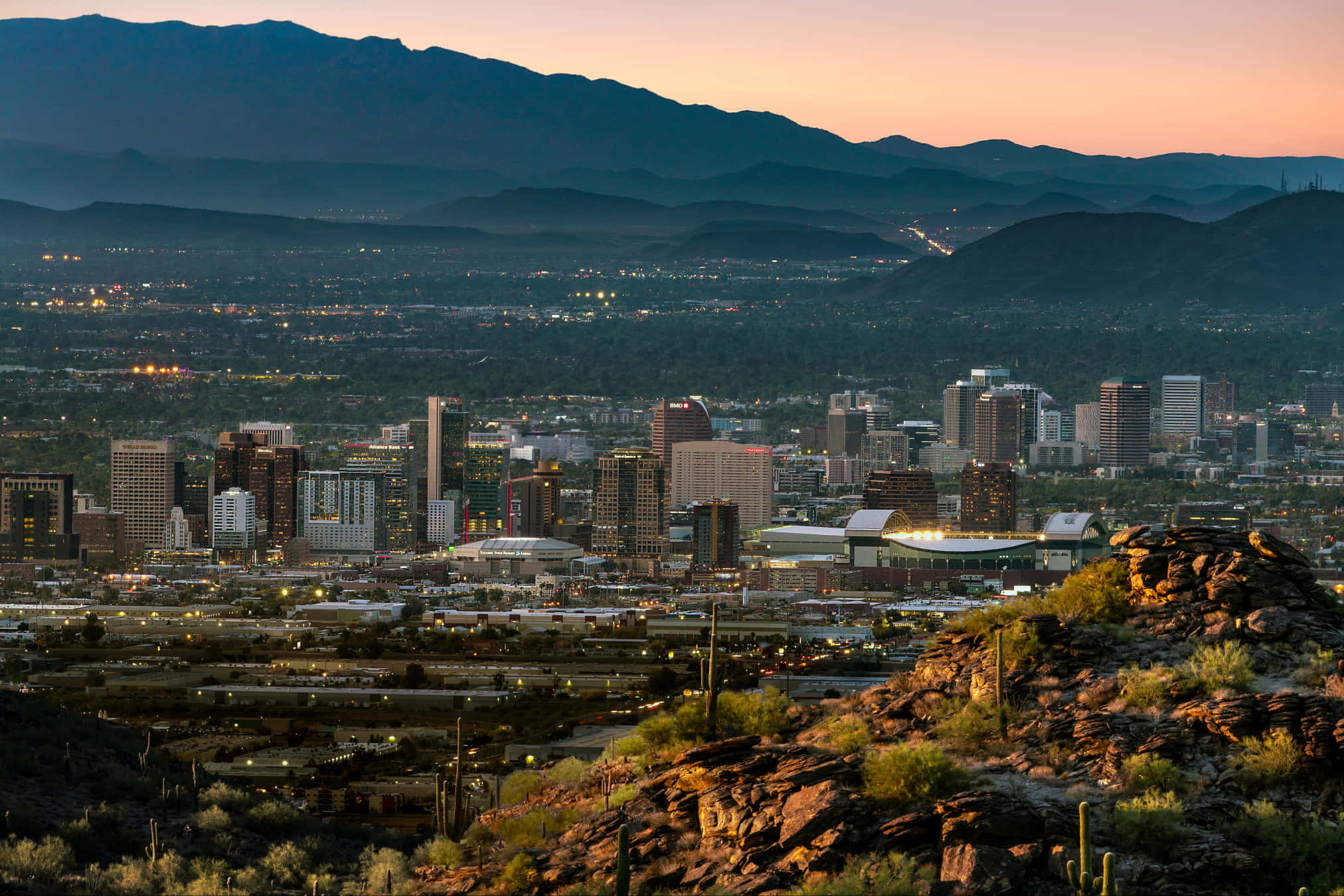 Phoenix Arizona 桌布 壁紙 — 在柔和的橙色天空下，沙漠丘陵與遠山之間，一片廣闊的城市景觀，摩天大樓在黃昏時分燈火通明。