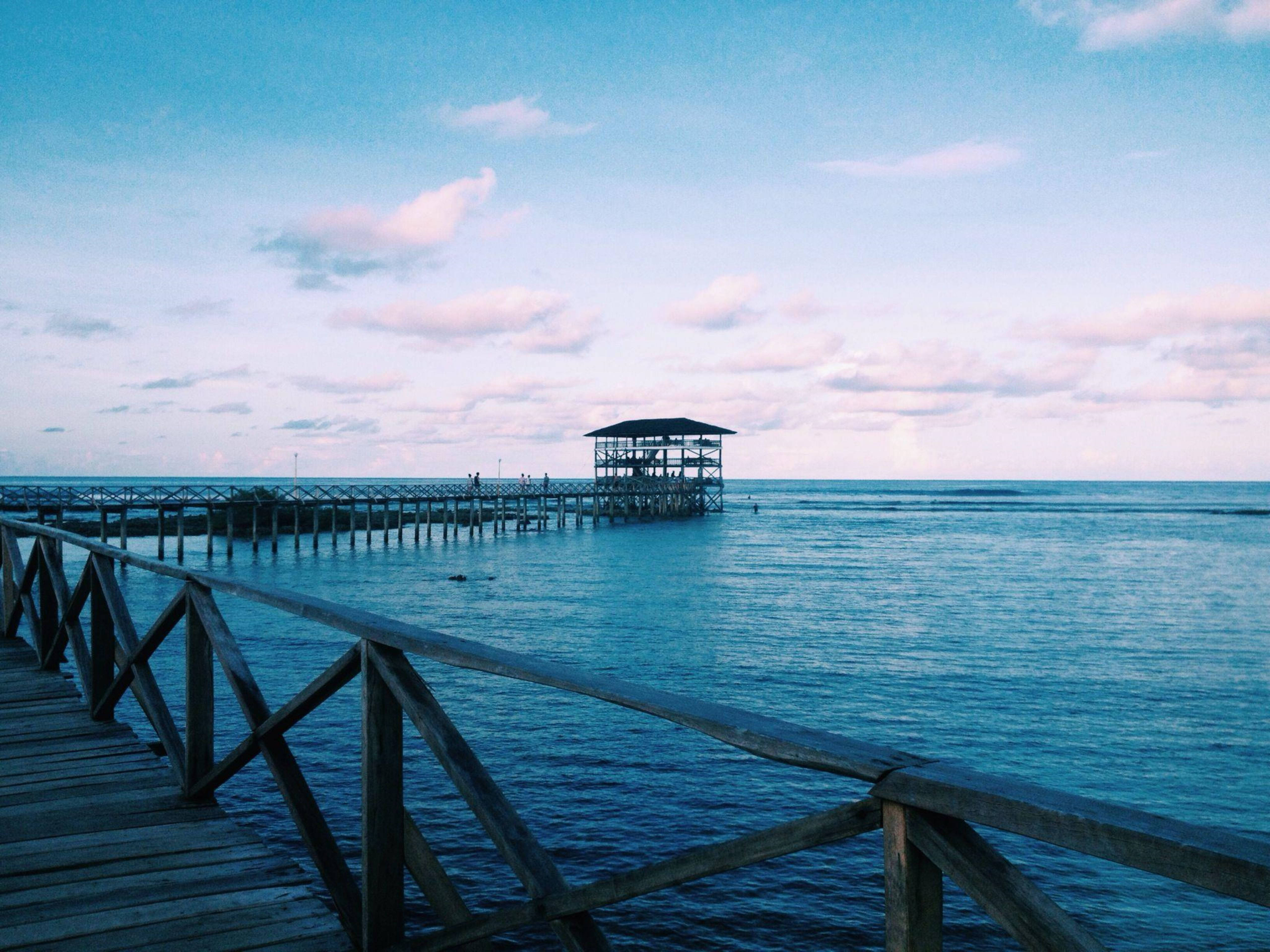 Siargao 桌布 壁紙 — 在柔和的粉彩天空下，黃昏時分，一個木製碼頭延伸到平靜的藍色海水中，天空中有散落的雲朵。