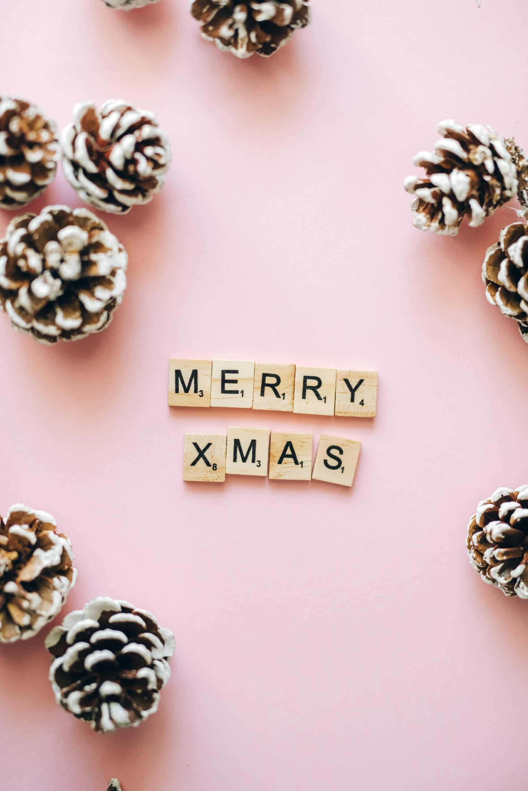 iPhone Christmas Aesthetic 桌布 壁紙 — 在柔和的粉紅色背景上，用木製瓷磚拼寫的「聖誕快樂」，周圍環繞著白雪覆蓋的松果。