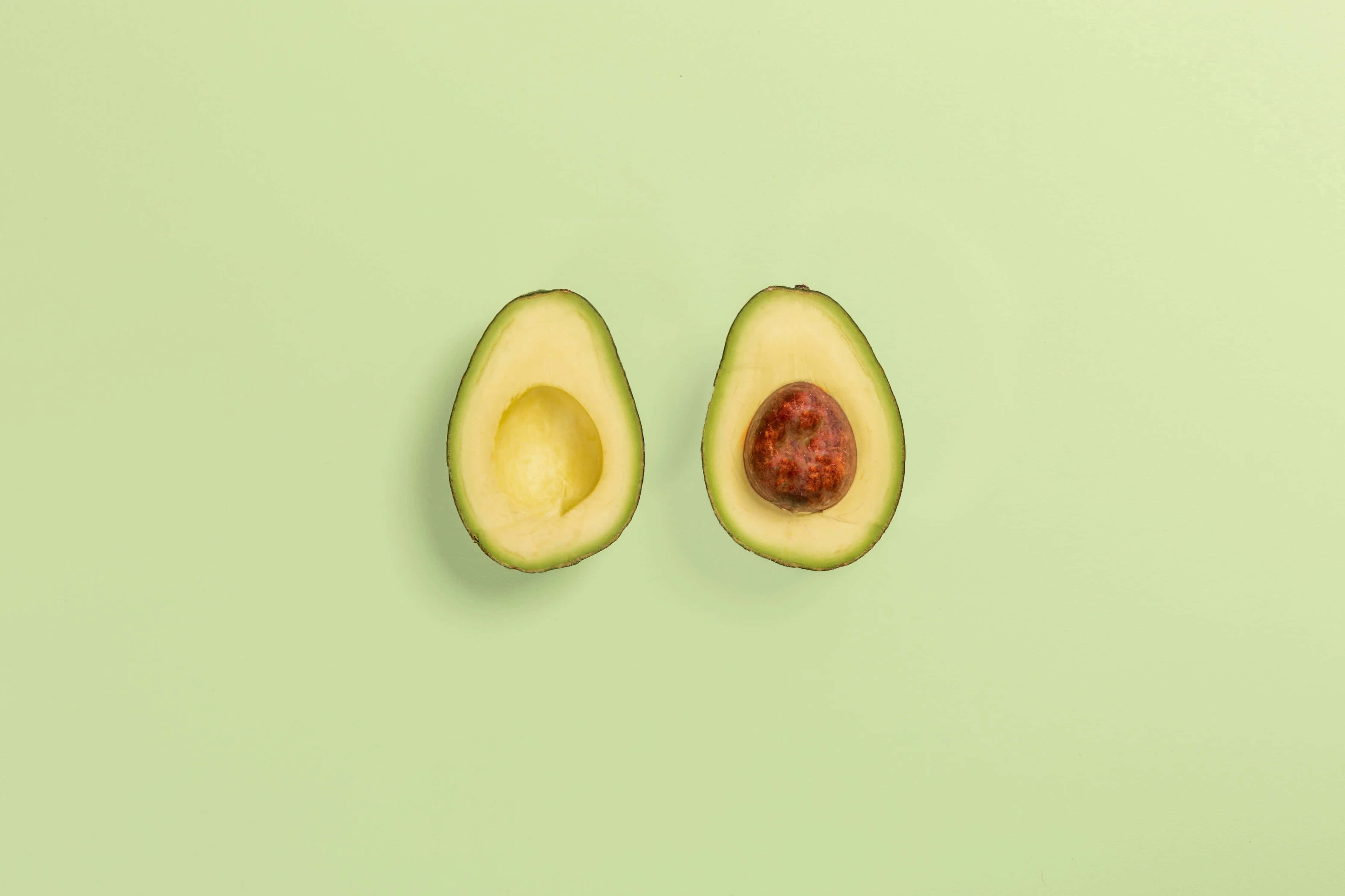Avocado 桌布 壁紙 — 在柔和的薄荷綠色背景上，一個成熟的酪梨被切成兩半，露出奶油狀的果肉和棕色的果核。