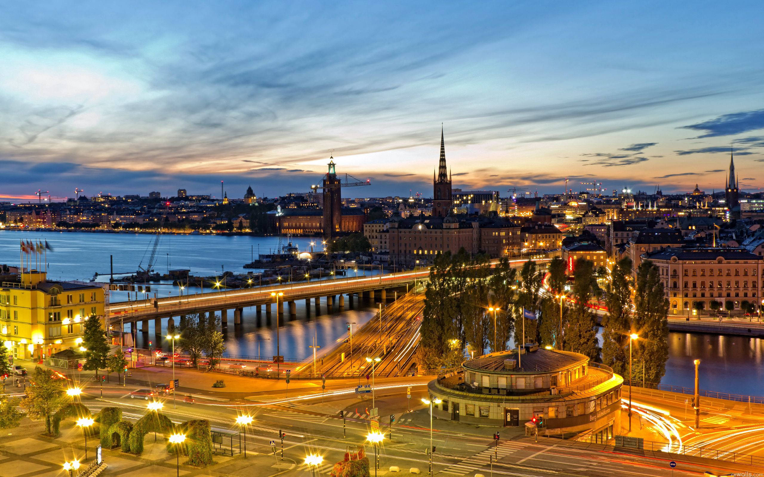 Sweden 桌布 壁紙 — 在柔和的藍天下，黃昏時分充滿活力的城市景觀，橋樑和歷史尖塔在平靜的水面上倒映著燈光。