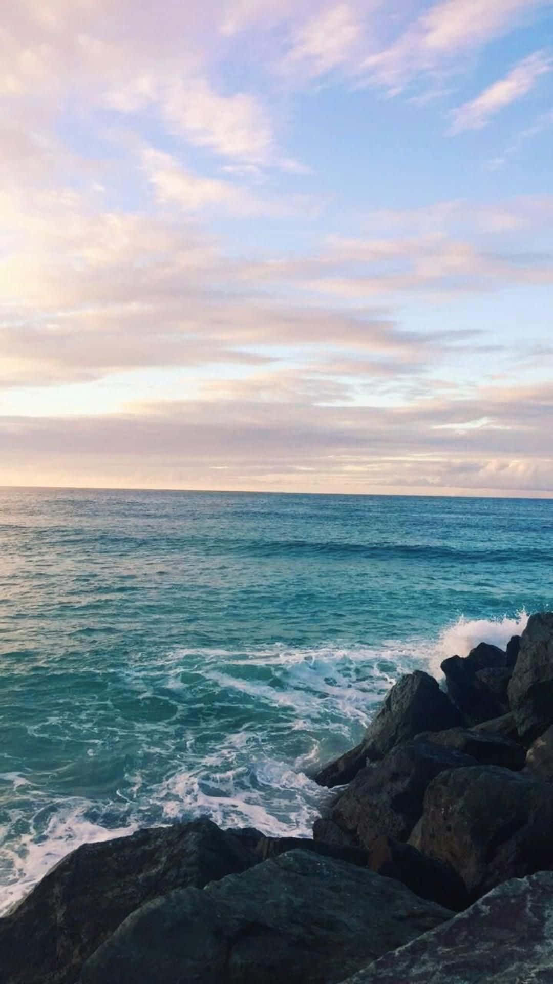 Ocean Aesthetic Phone 桌布 壁紙 — 在柔和粉彩日落天空和縷縷雲彩下，綠松石色的海浪拍打著深色岩石。