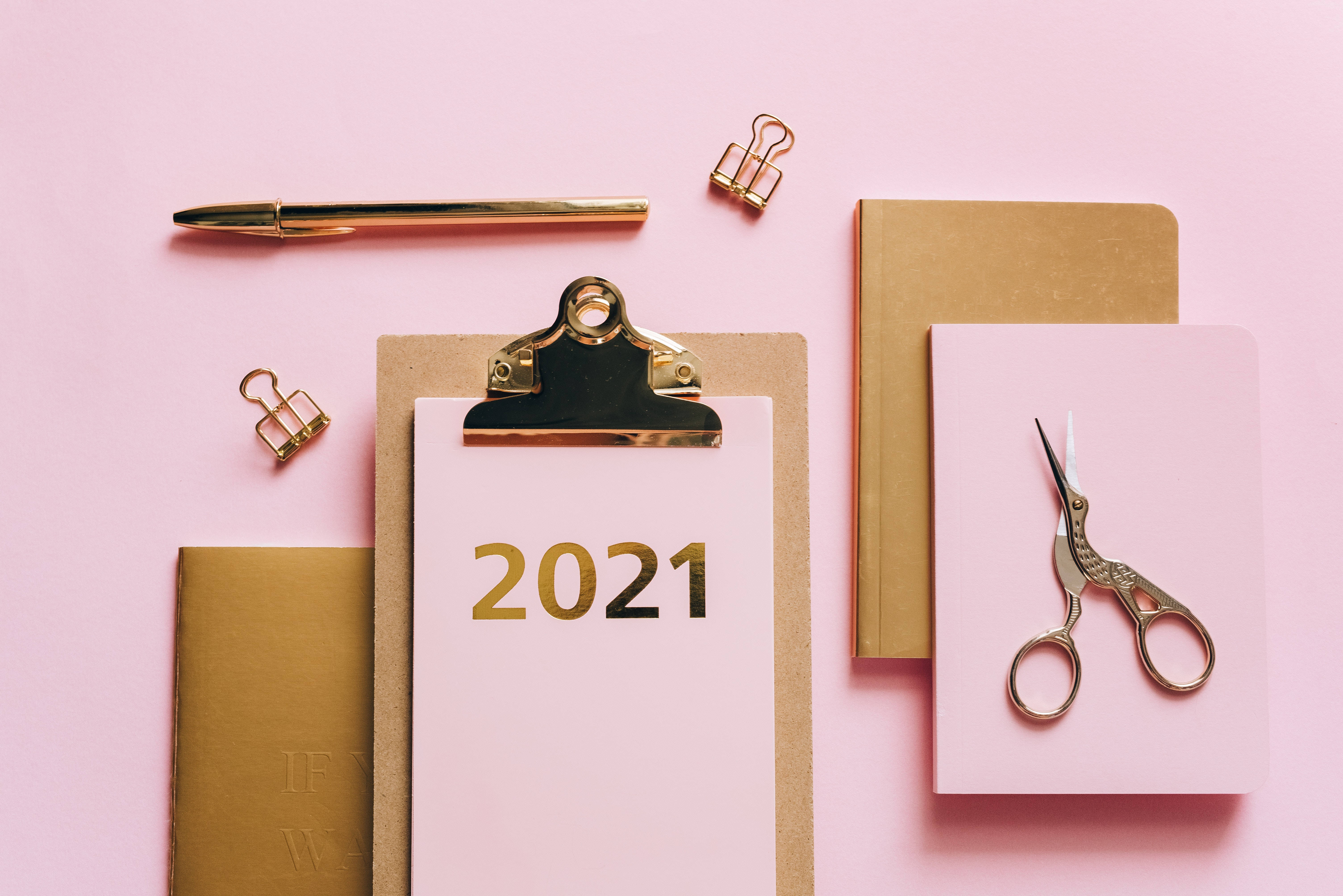 2021 桌面 桌布 壁紙 — 在柔軟的粉紅色背景上平鋪金色辦公用品和筆記本，剪貼板上印有「2021」。