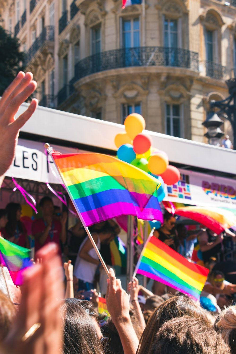 Pride Flag 桌布 壁紙 — 在歐洲城市街道上，陽光明媚的日間遊行中，一群充滿活力的人群揮舞著彩虹旗和彩色氣球。