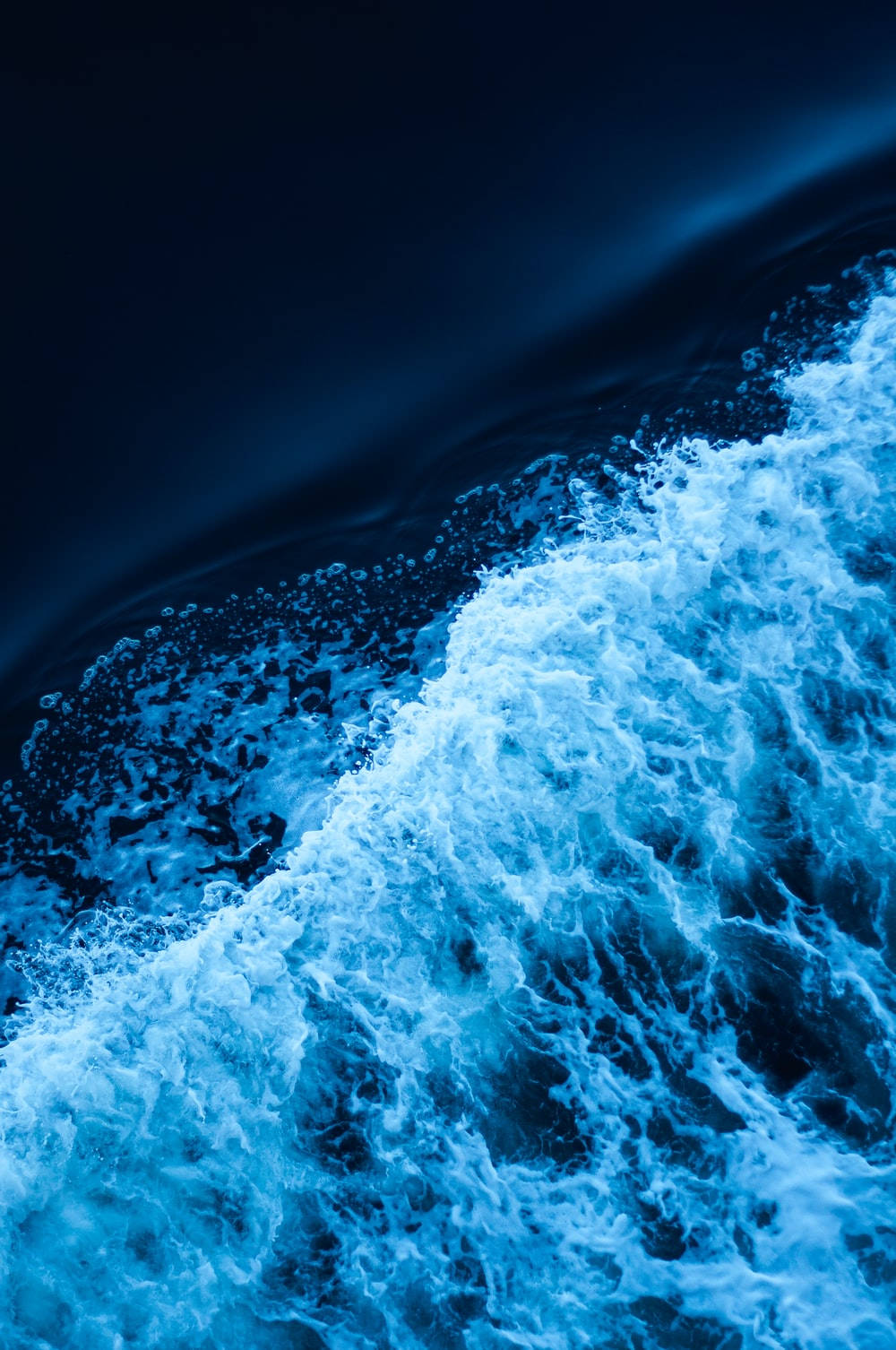 Aesthetic Dark Blue 桌布 壁紙 — 在氣氛燈光下，泡沫海浪猛烈衝擊深藍色海水的戲劇性特寫。