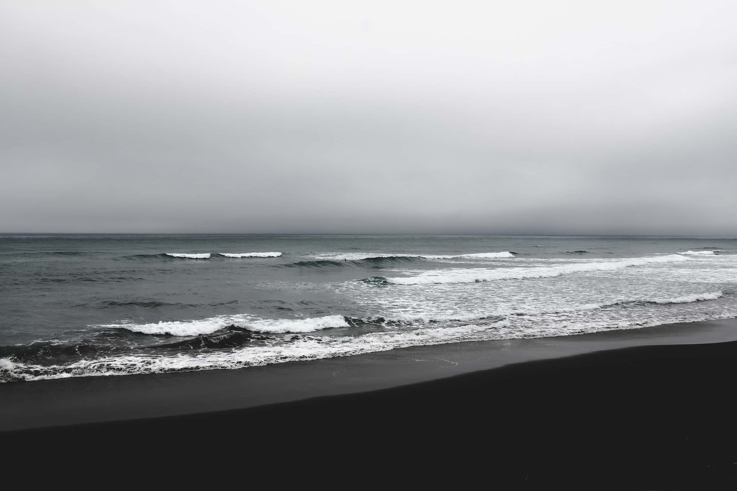 Dark Ocean 桌布 壁紙 — 在沉重的灰色天空下，海浪拍打著黑色的火山沙灘，營造出陰鬱的海岸場景。