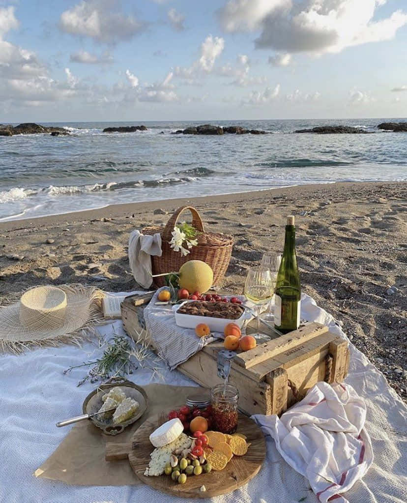 Beach Picnic 桌布 壁紙 — 在沙灘上展開一場奢華的海灘野餐，木箱裡裝滿了起司、水果、葡萄酒，旁邊還有一個編織籃，伴隨著海浪拍打，天空部分多雲。