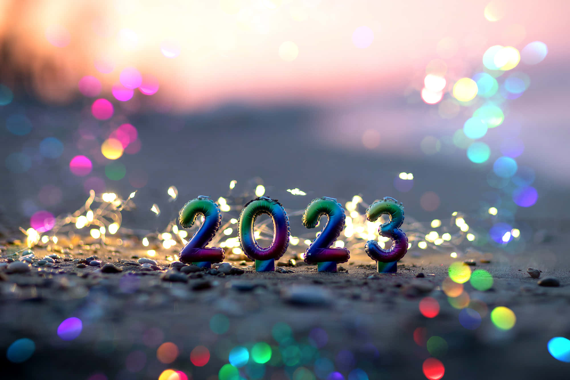 新年 桌布 壁紙 — 在沙灘上閃爍燈光和日落散景背景下的2023年彩虹色氣球數字。