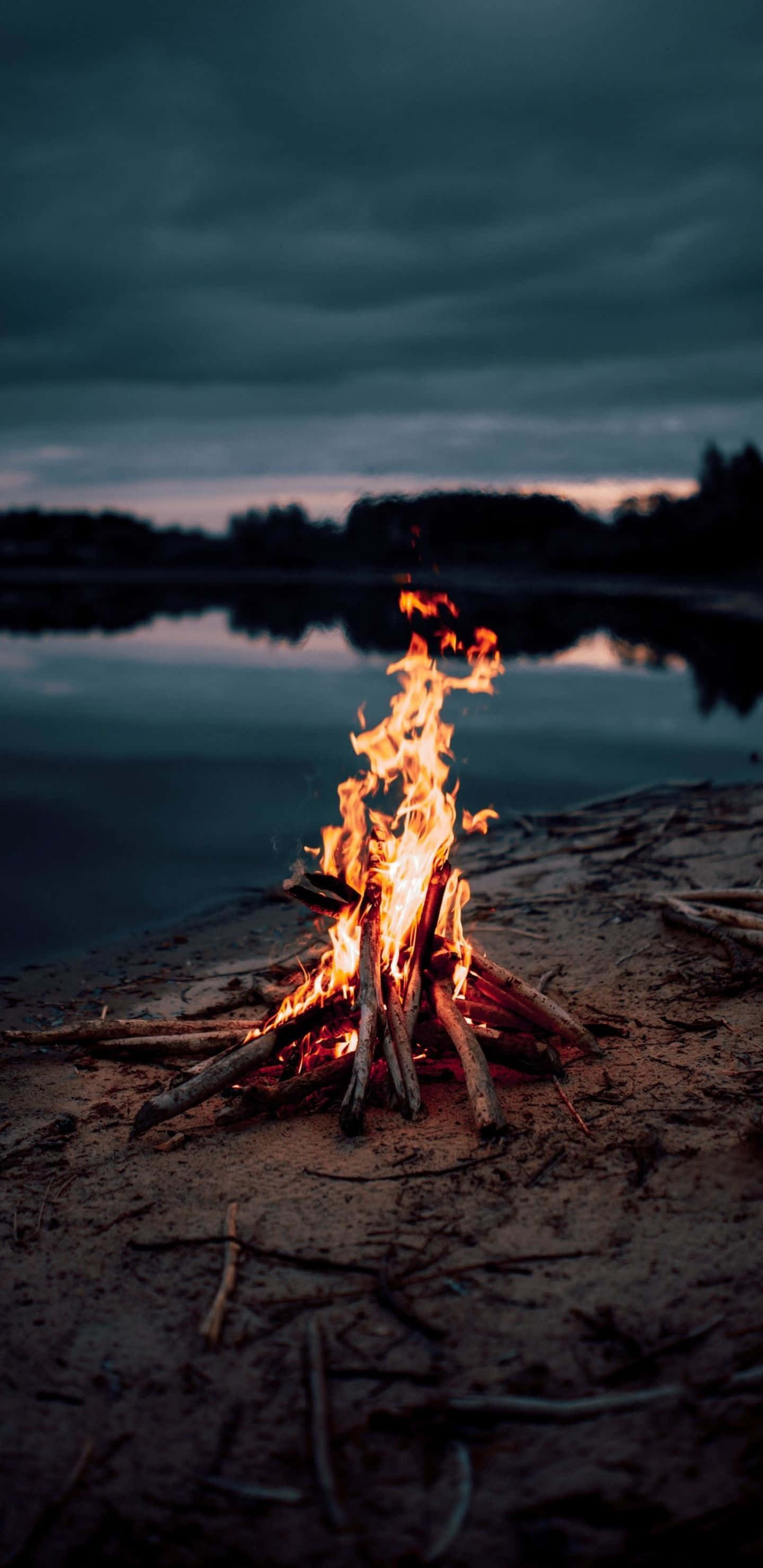 Beach Bonfire 桌布 壁紙 — 在沙質湖岸上，一堆熊熊燃燒的營火在陰鬱的暮色天空下明亮地燃燒，黑暗的雲朵和遠處的樹木倒映在平靜的水面上。