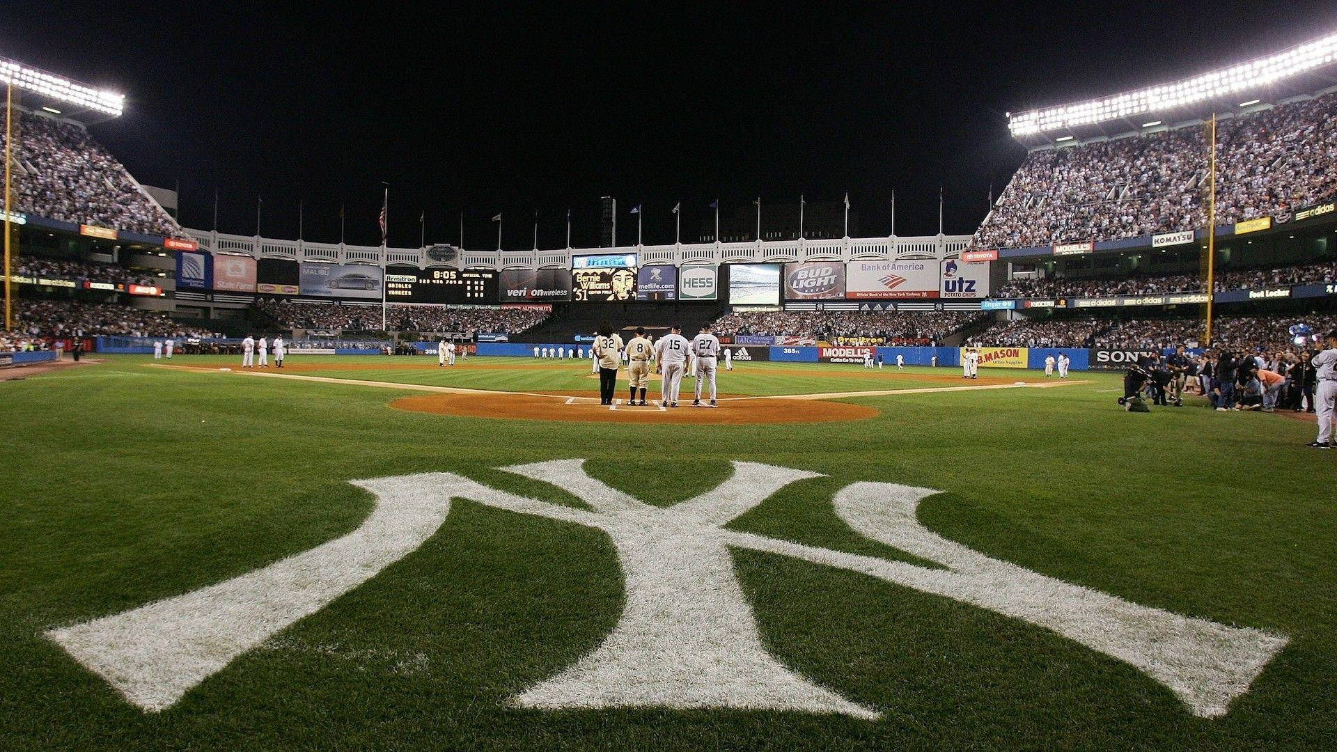 Yankees Logo 桌布 壁紙 — 在洋基球場舉行的夜間棒球比賽，球員站在標誌性的NY標誌上，球場燈光璀璨。